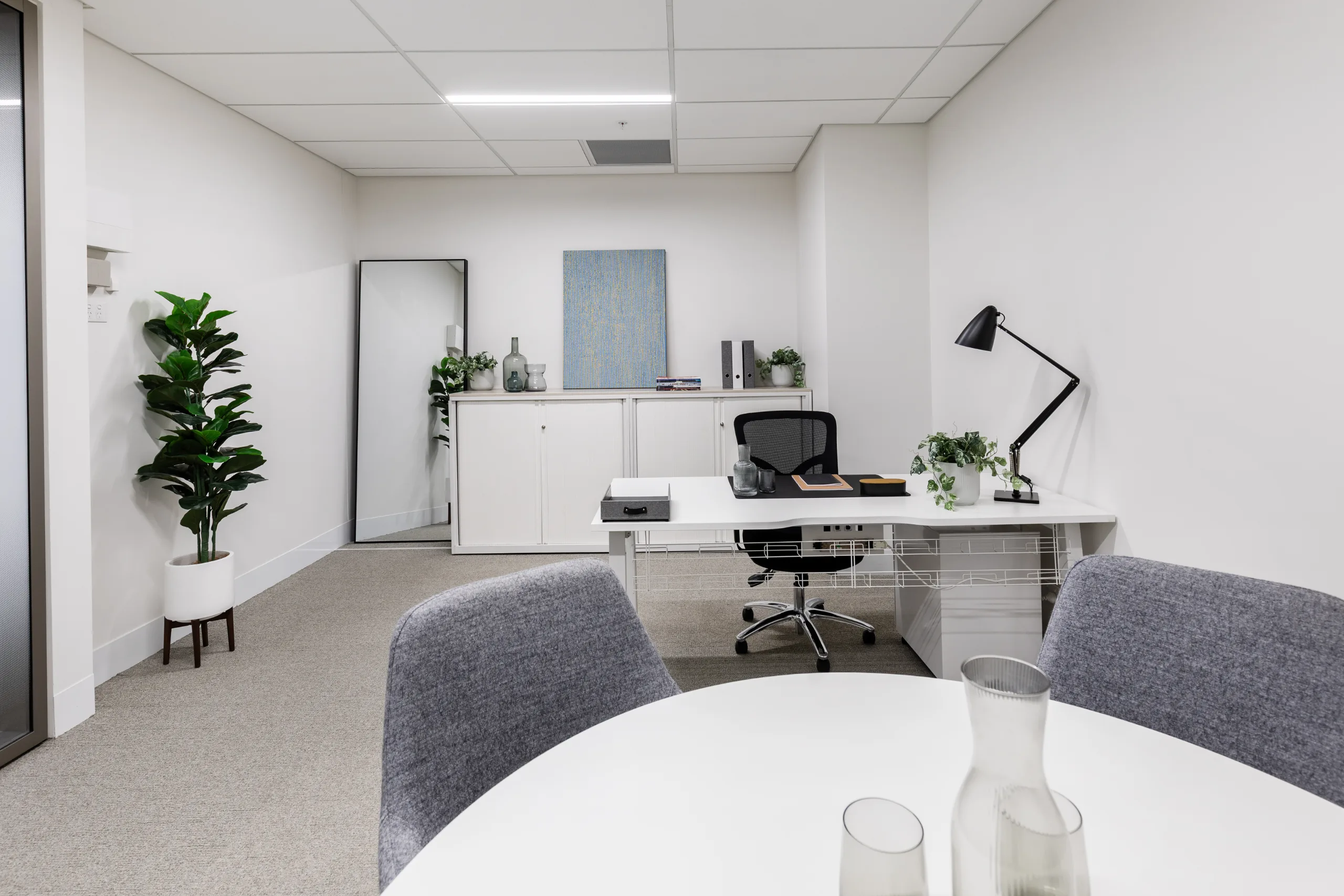 Brightly lit office space featuring a white desk, black ergonomic chair, and a potted plant against white walls.