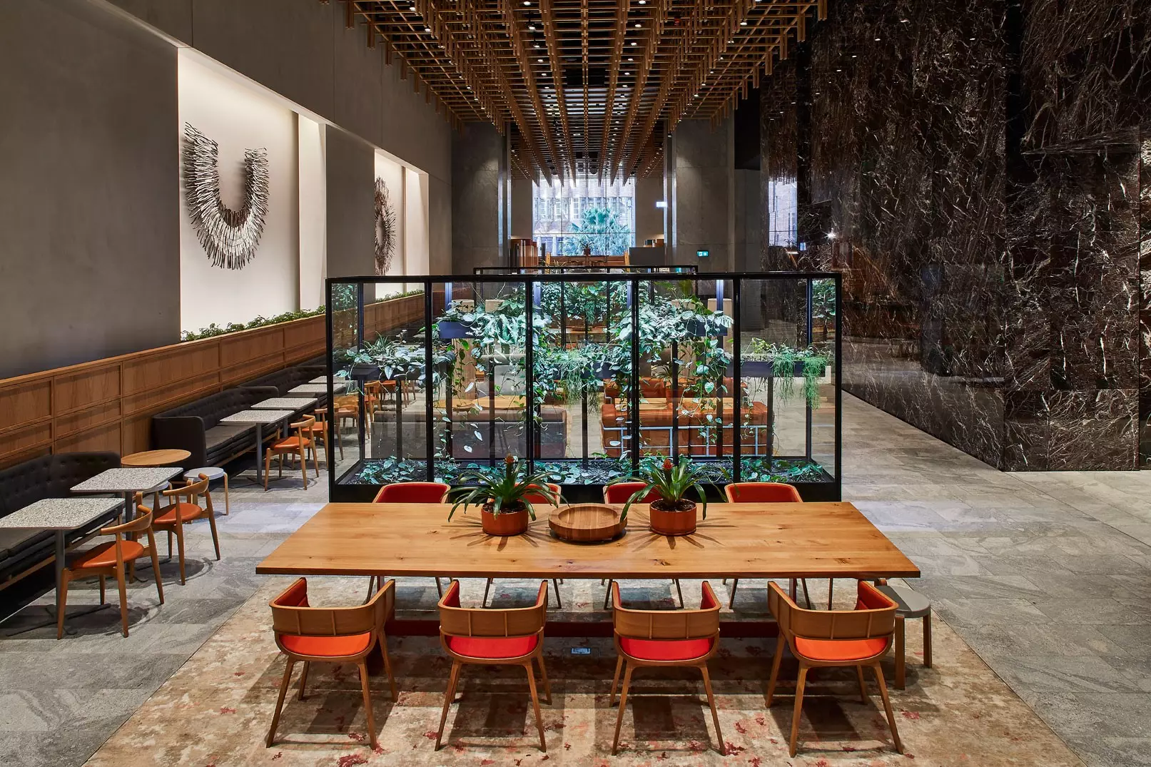 Modern office lounge featuring a long wooden communal table, indoor greenery, and warm ambient lighting.