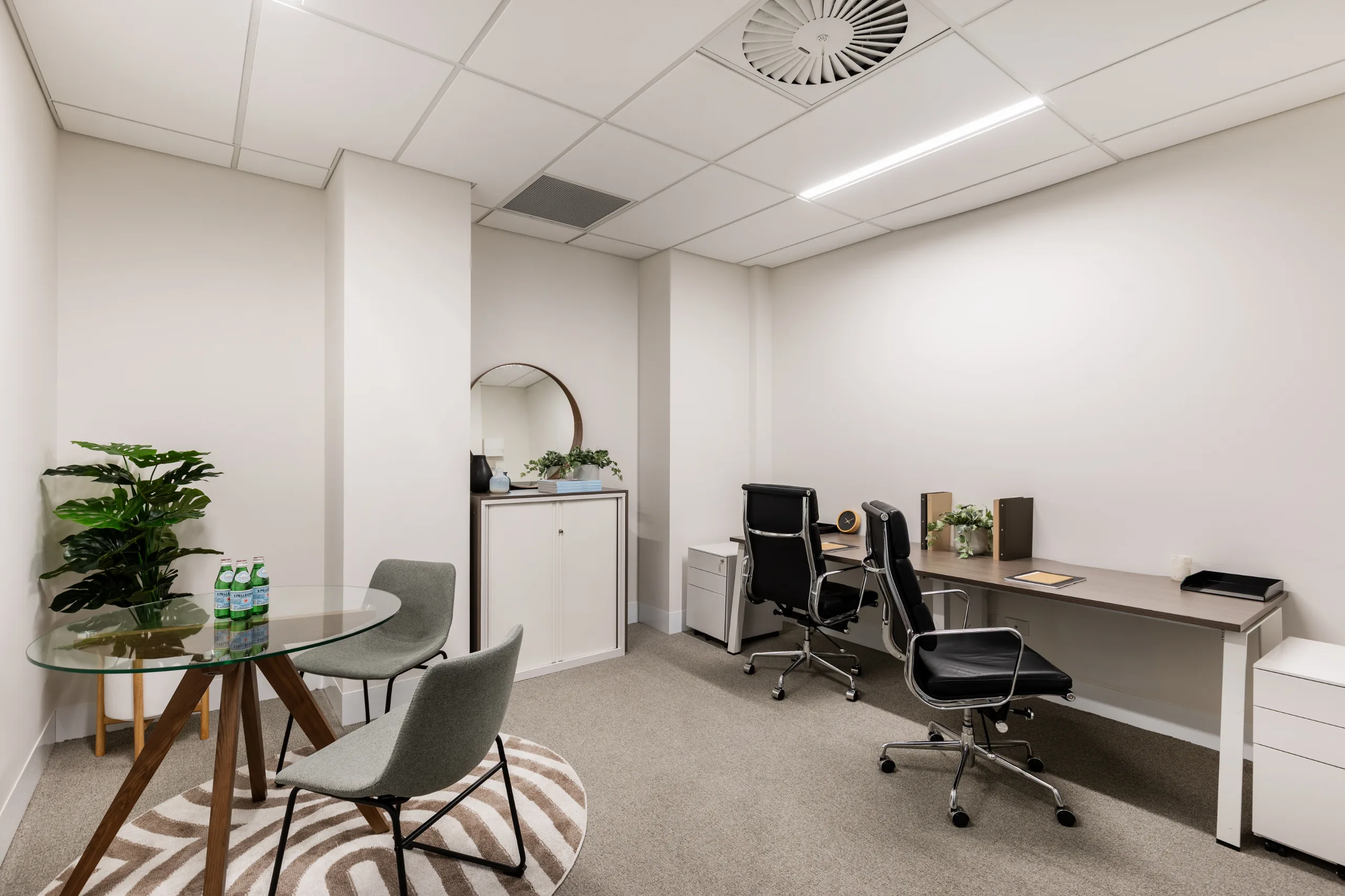 Bright office space featuring a glass meeting table, grey chairs, and a long desk with black office chairs.