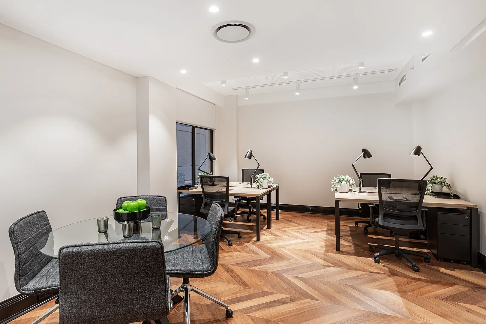 Bright office space featuring wooden desks, black ergonomic chairs, and a glass meeting table on herringbone flooring.