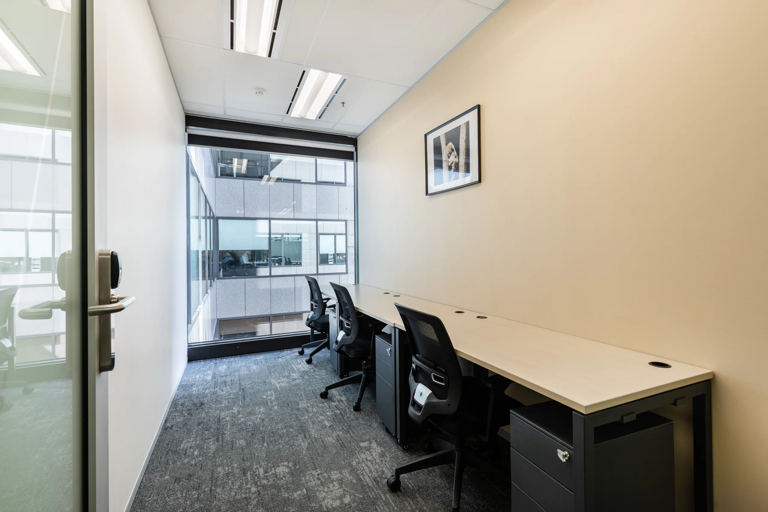 Bright office room featuring a long white desk, black chairs, and large windows overlooking an adjacent building.