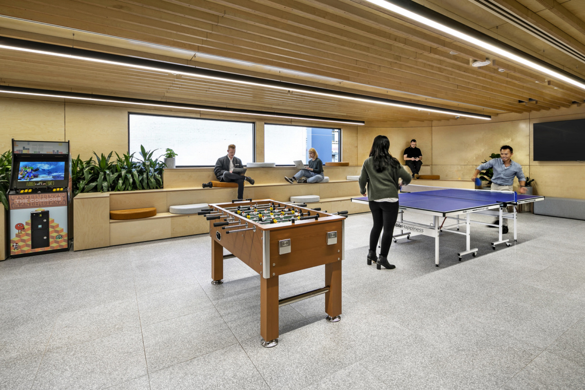 Modern office breakroom featuring a foosball table, ping pong table, arcade machine, and people relaxing on wooden seating.