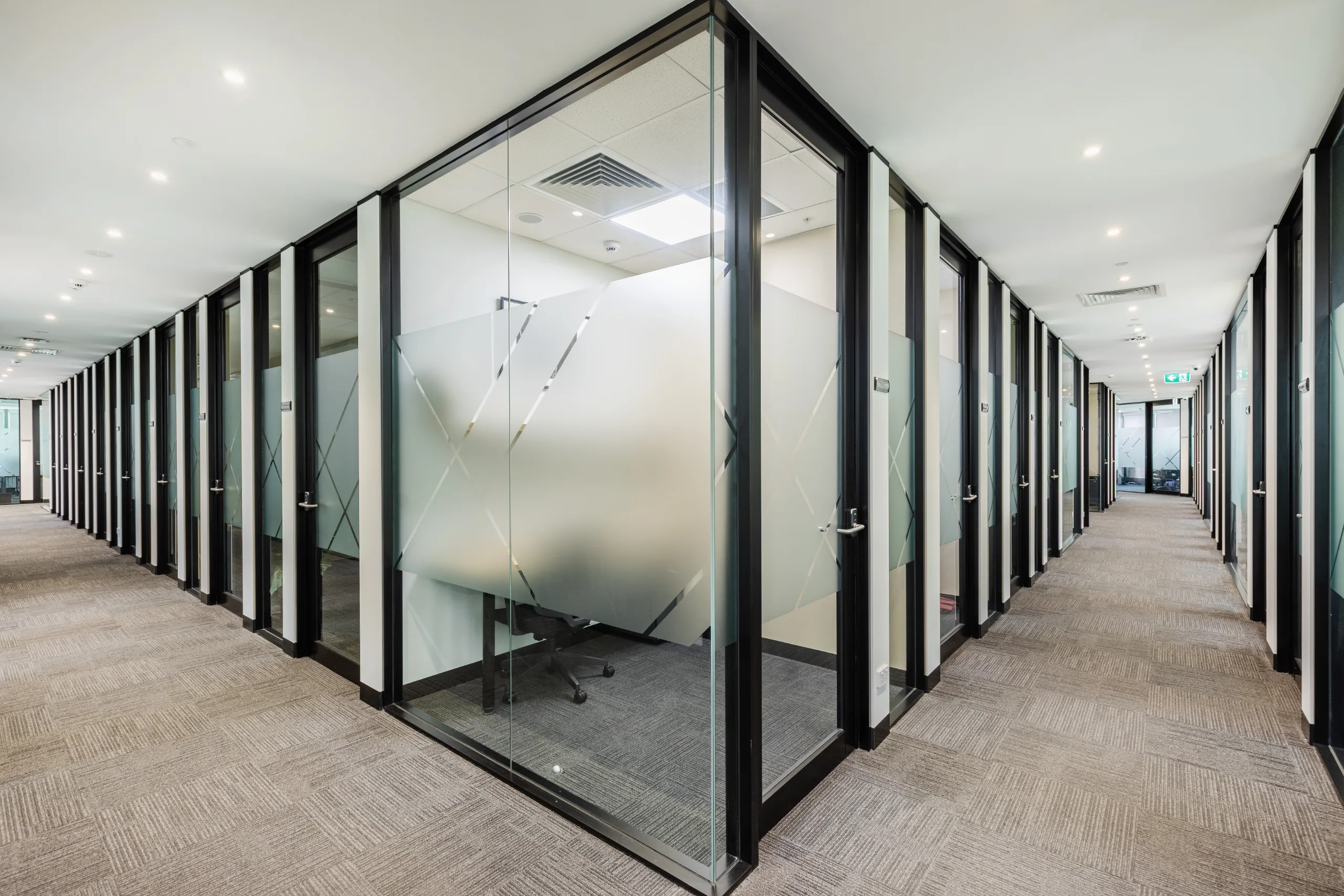 A modern office corridor features glass-walled private meeting rooms with frosted privacy patterns and grey carpeting.