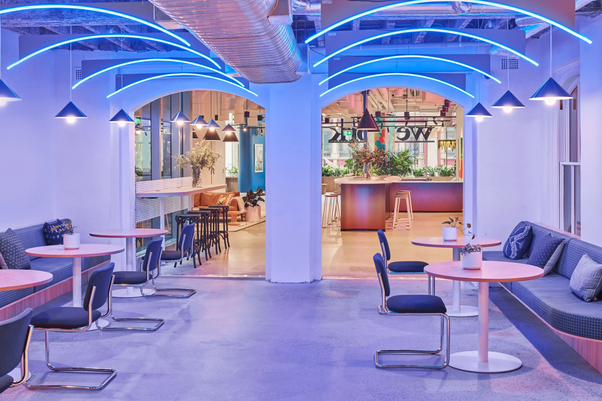 Modern office lounge with blue neon ceiling lights, circular tables, and cushioned bench seating.