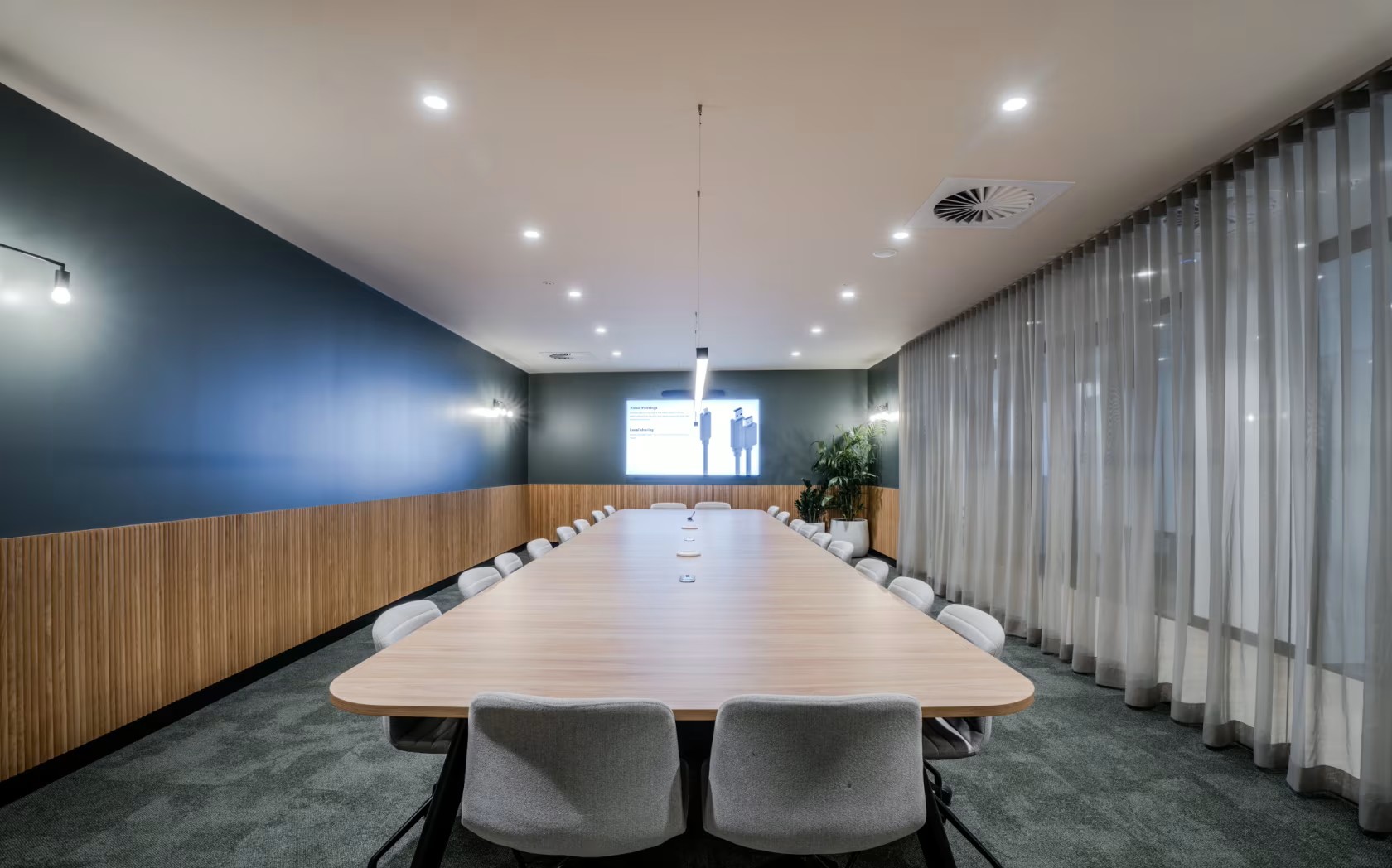 Modern conference room with a long wooden table, grey chairs, and large windows with sheer curtains.