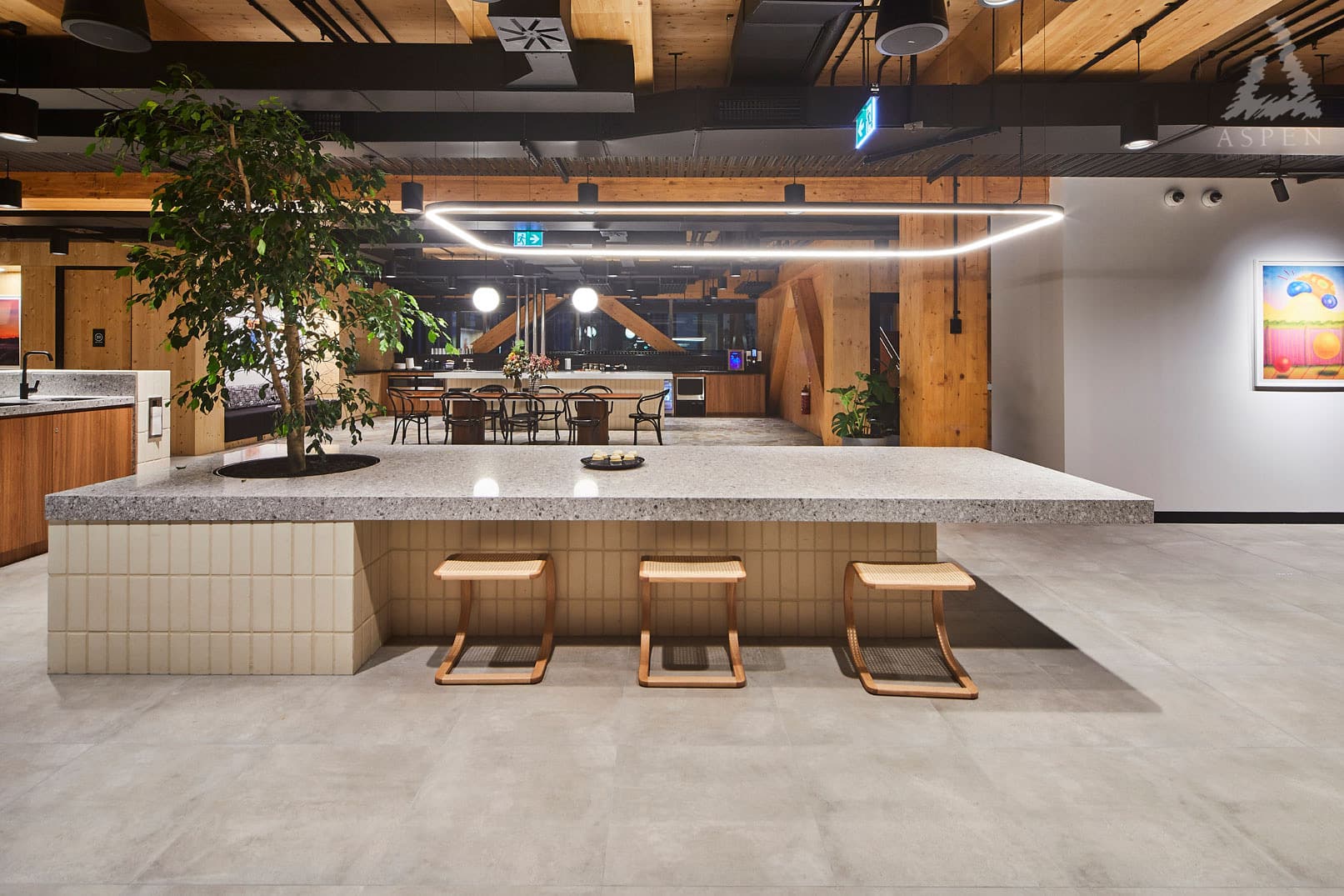 Modern office kitchen area with a long stone island, wooden stools, and a large indoor tree.
