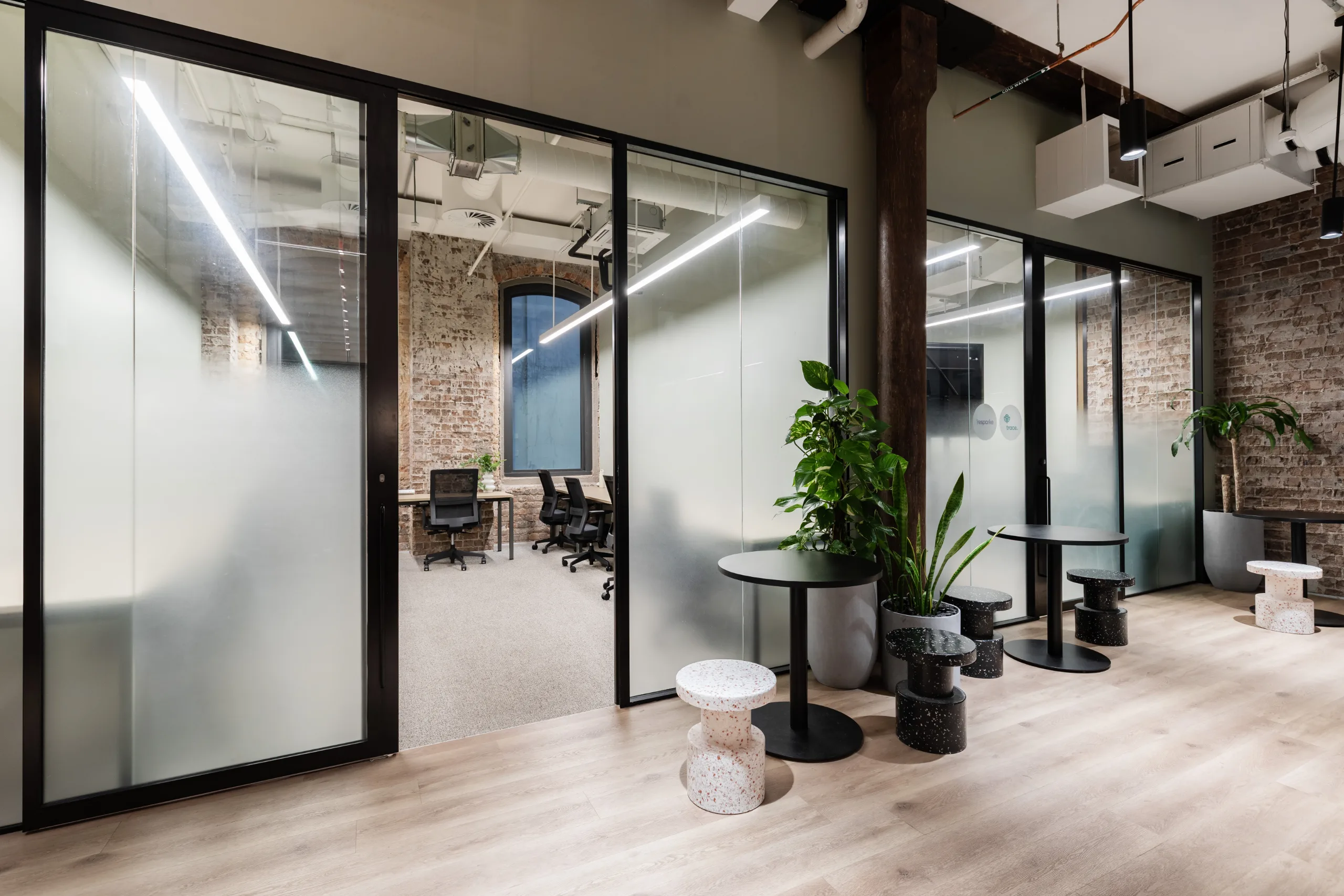 Modern office space featuring glass-walled meeting rooms, wooden floors, and small round tables with stools.
