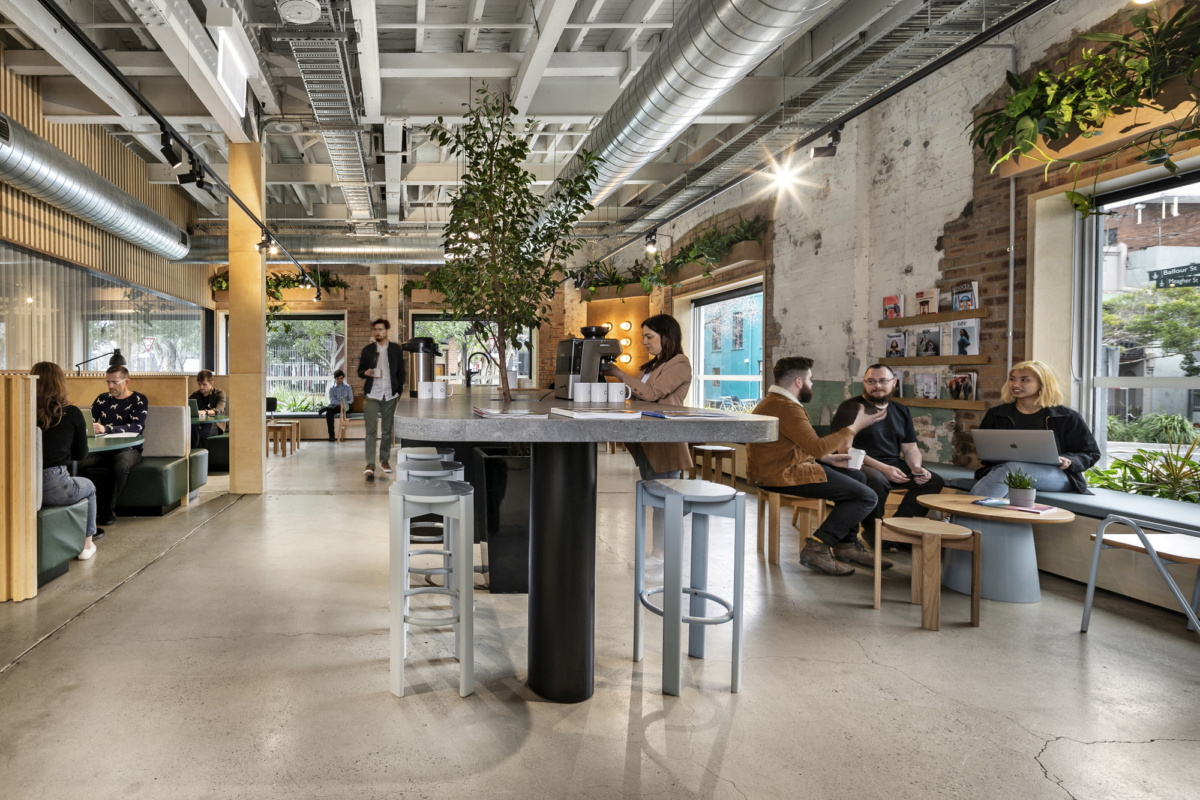 Bright, open-plan office space with high ceilings, industrial features, and people sitting at communal tables and lounge areas.
