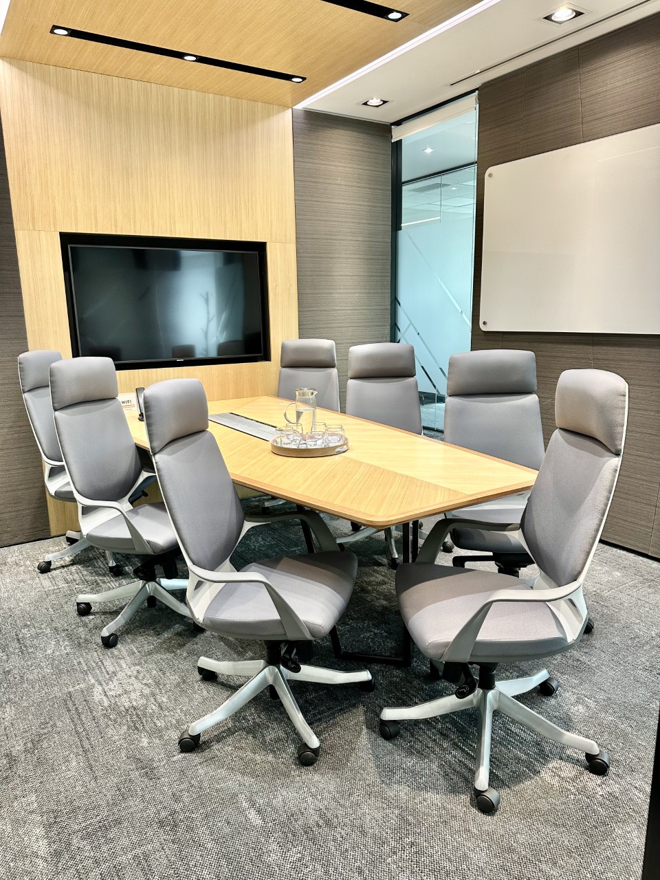 Modern meeting room featuring a light wood table, grey ergonomic chairs, and a large wall-mounted screen.