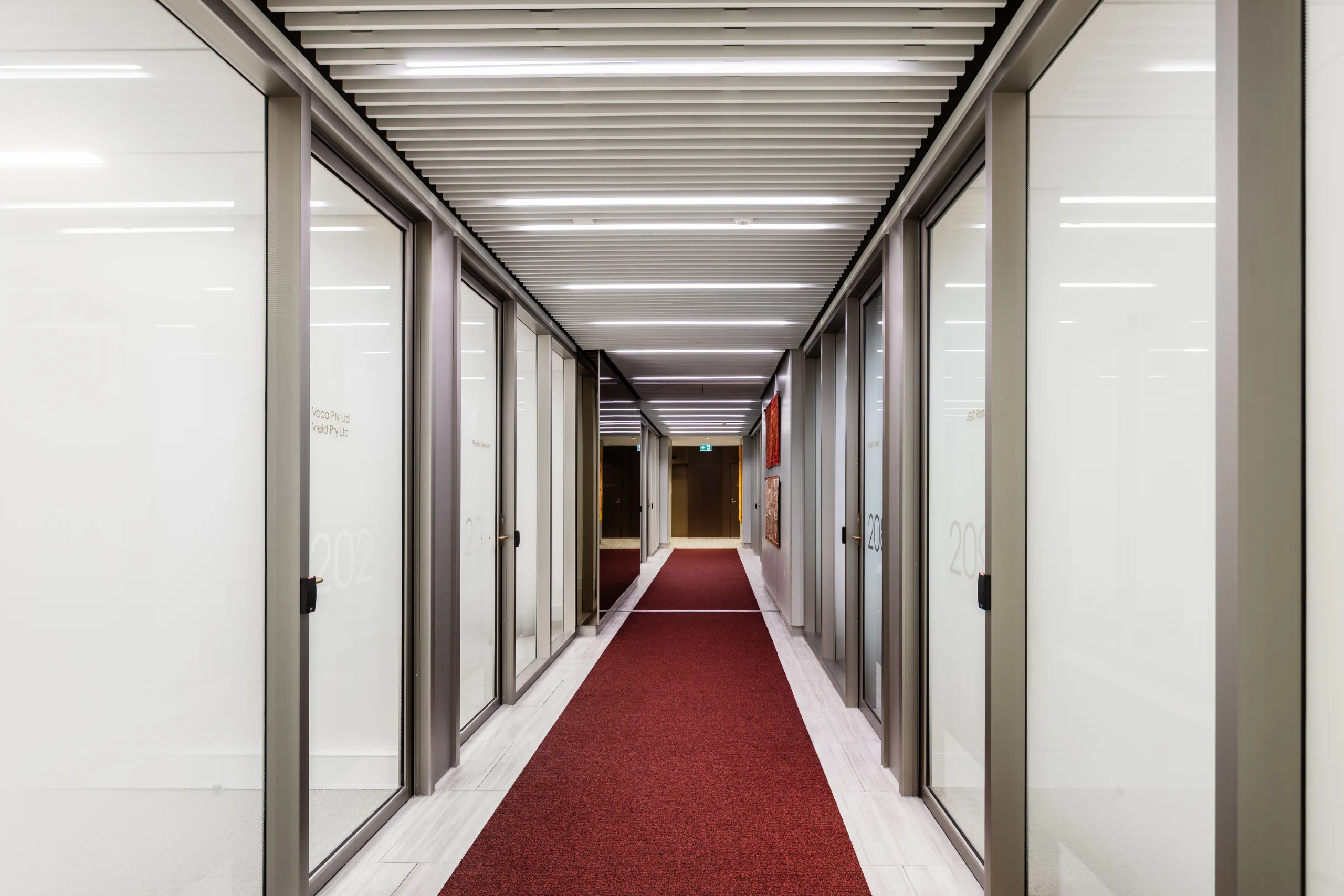 A long office corridor with frosted glass doors, white walls, and a red carpet runner.