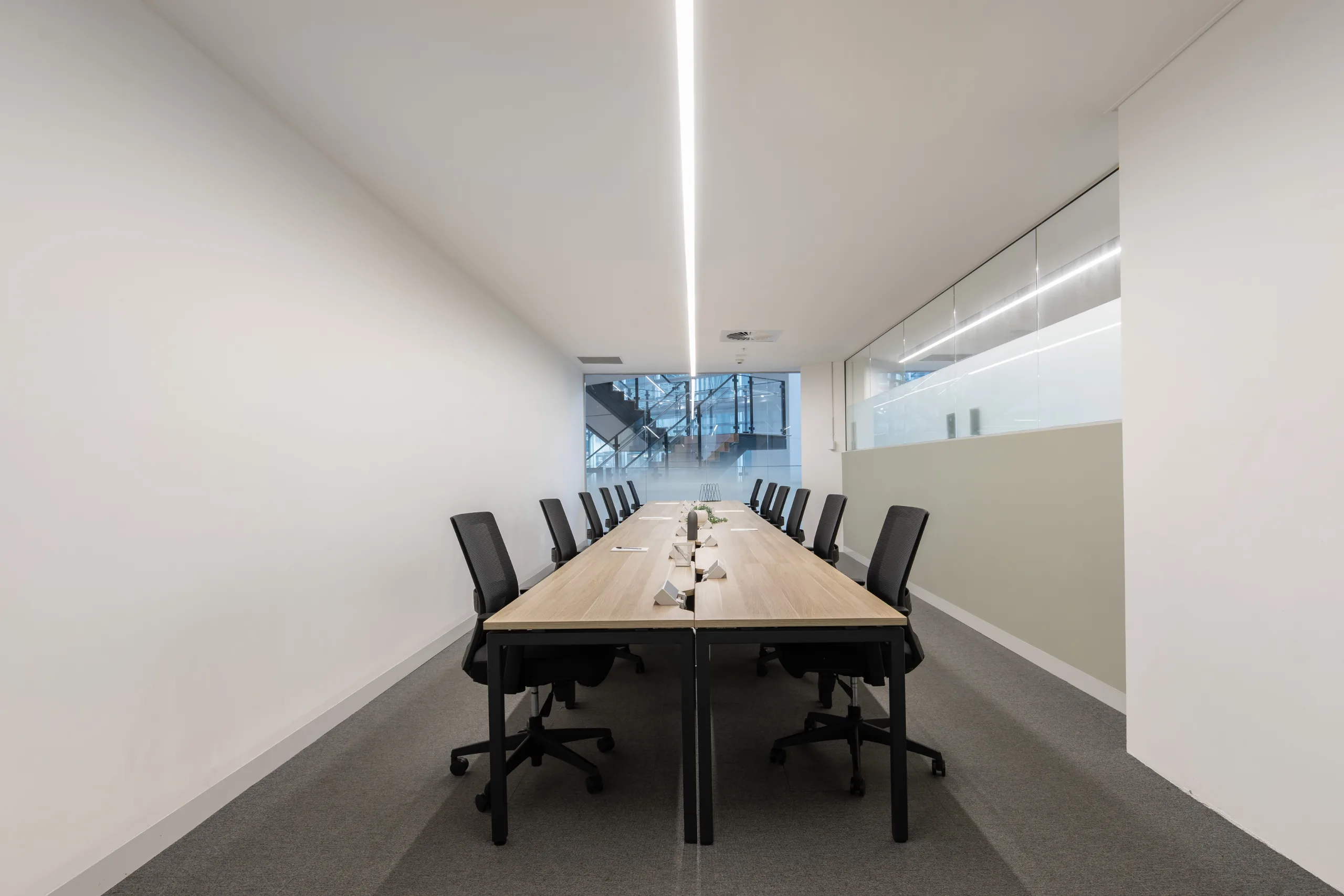 Long wooden meeting table with black ergonomic chairs in a bright, modern office with large windows.