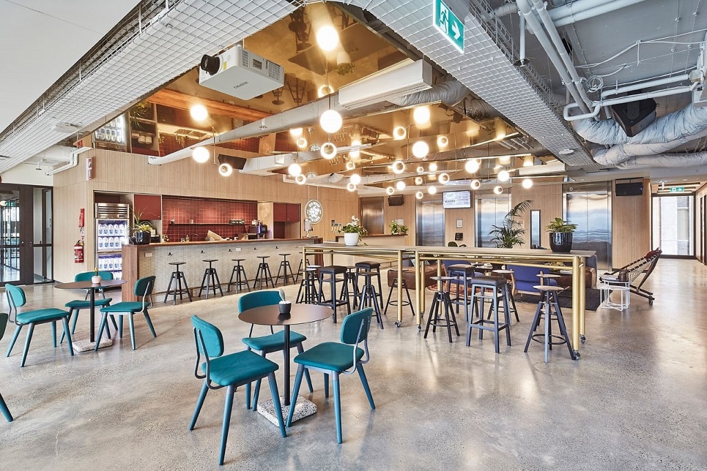 Modern open-plan office lounge featuring teal chairs, high bar seating, and bright industrial lighting.