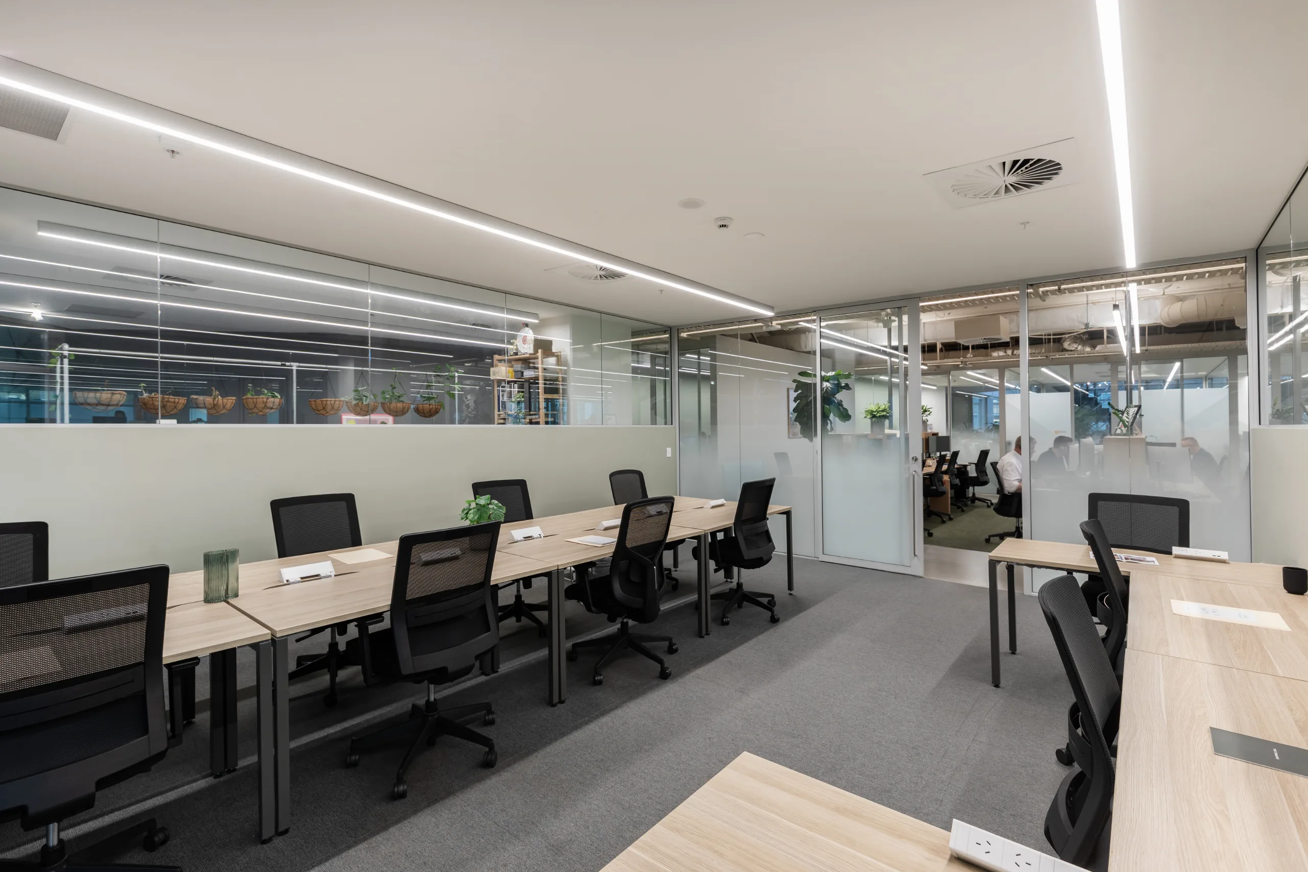 Modern open-plan office with long wooden desks, black ergonomic chairs, and bright integrated ceiling lighting.