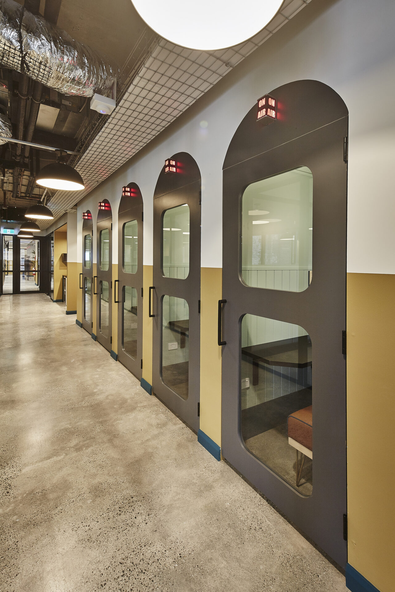 A row of modern grey private office pods with glass windows along a polished concrete hallway.