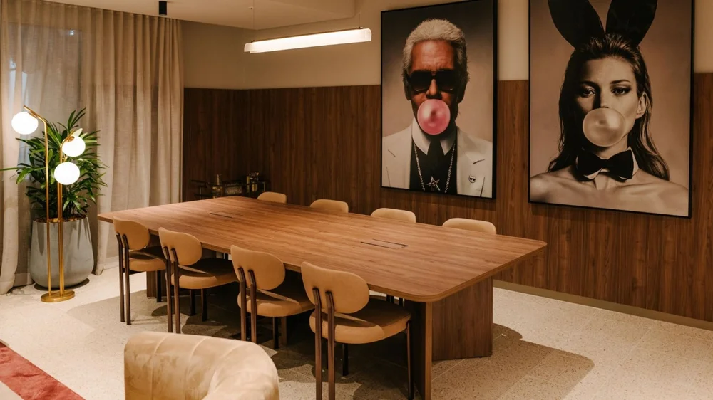 Modern meeting room featuring a long wooden table, tan chairs, and large portrait artworks on wood-panelled walls.