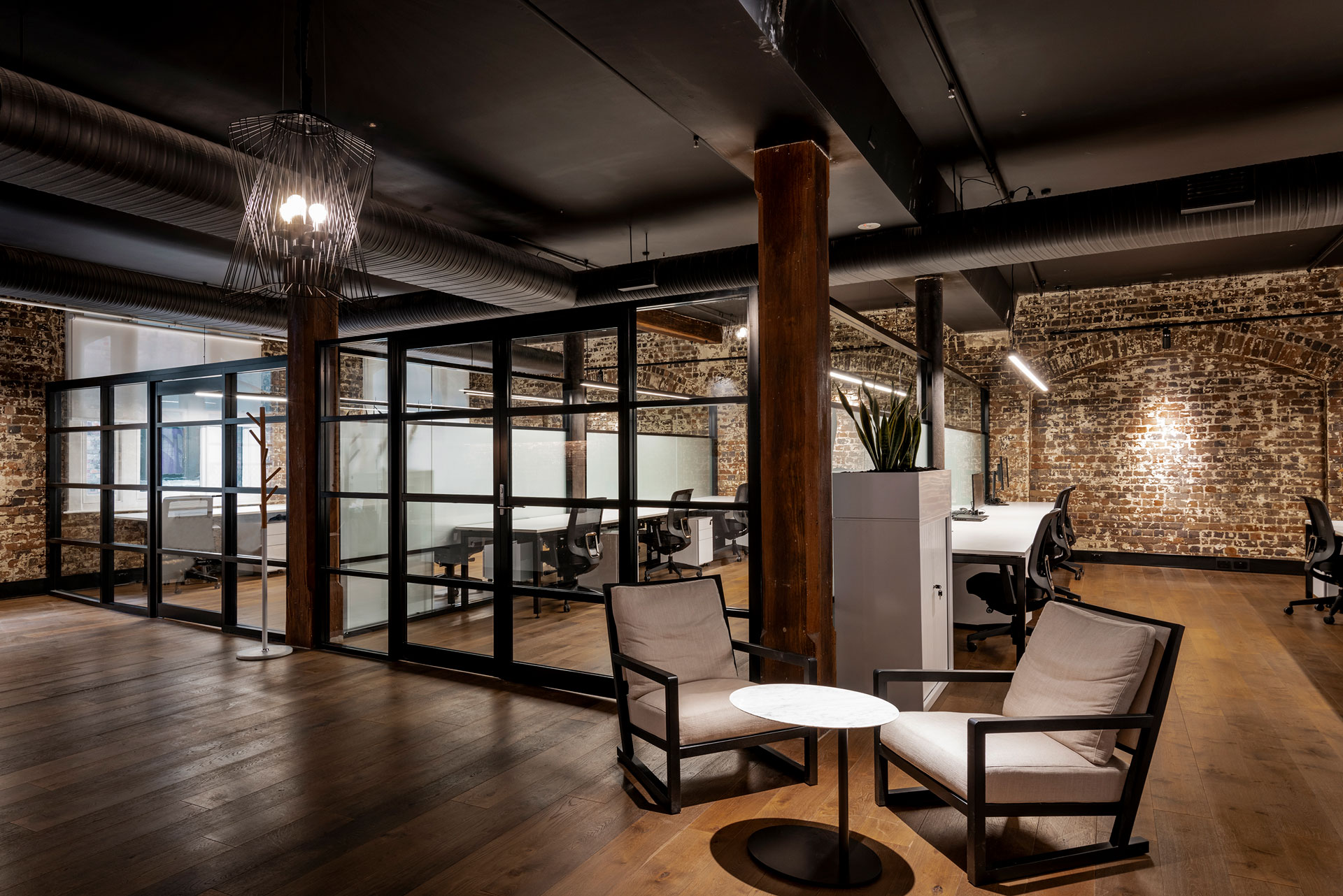 Modern open-plan office with glass partitions, wooden floors, exposed brick walls, and warm ambient lighting.