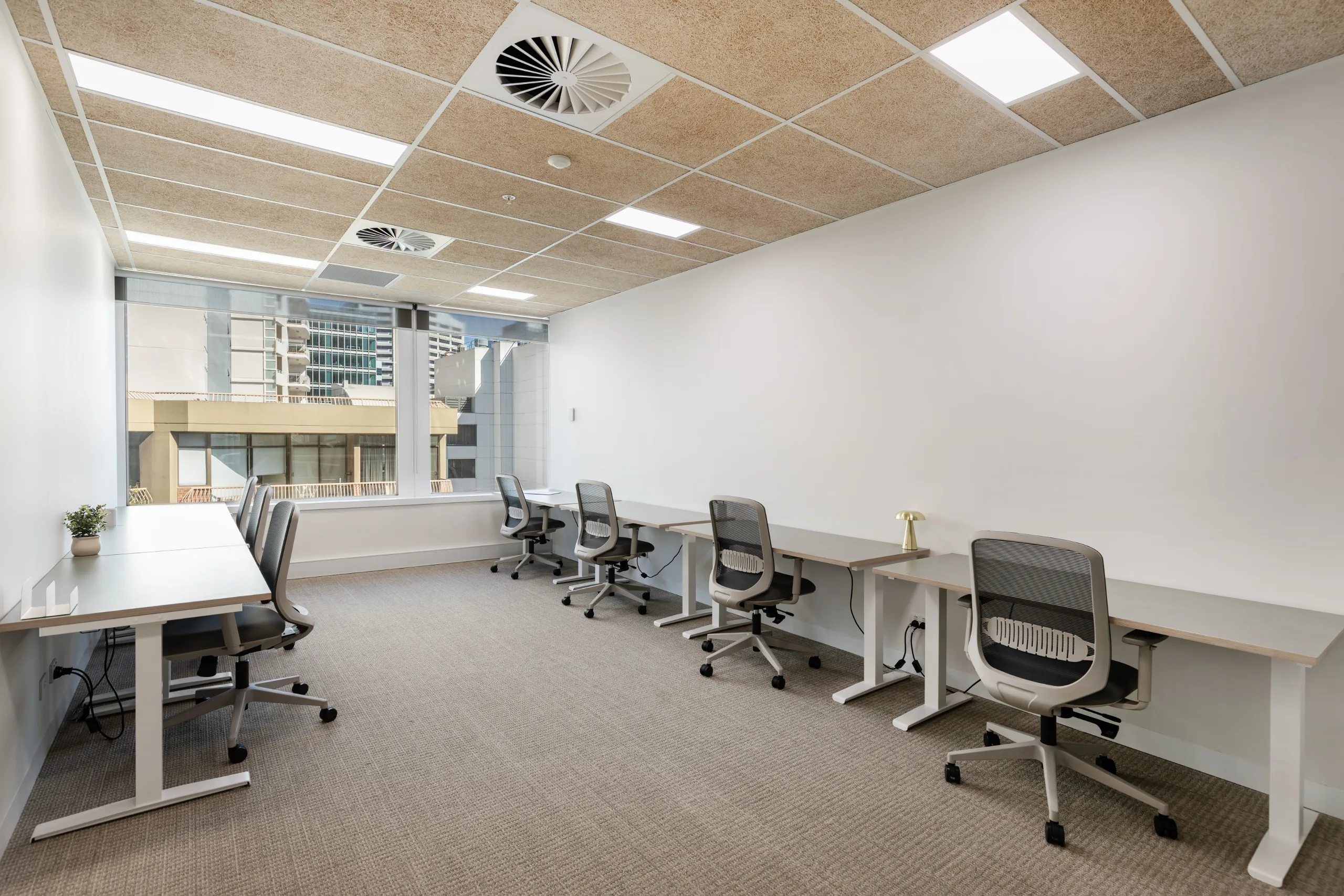 Bright office space with long white desks, ergonomic chairs, and large windows overlooking city buildings.