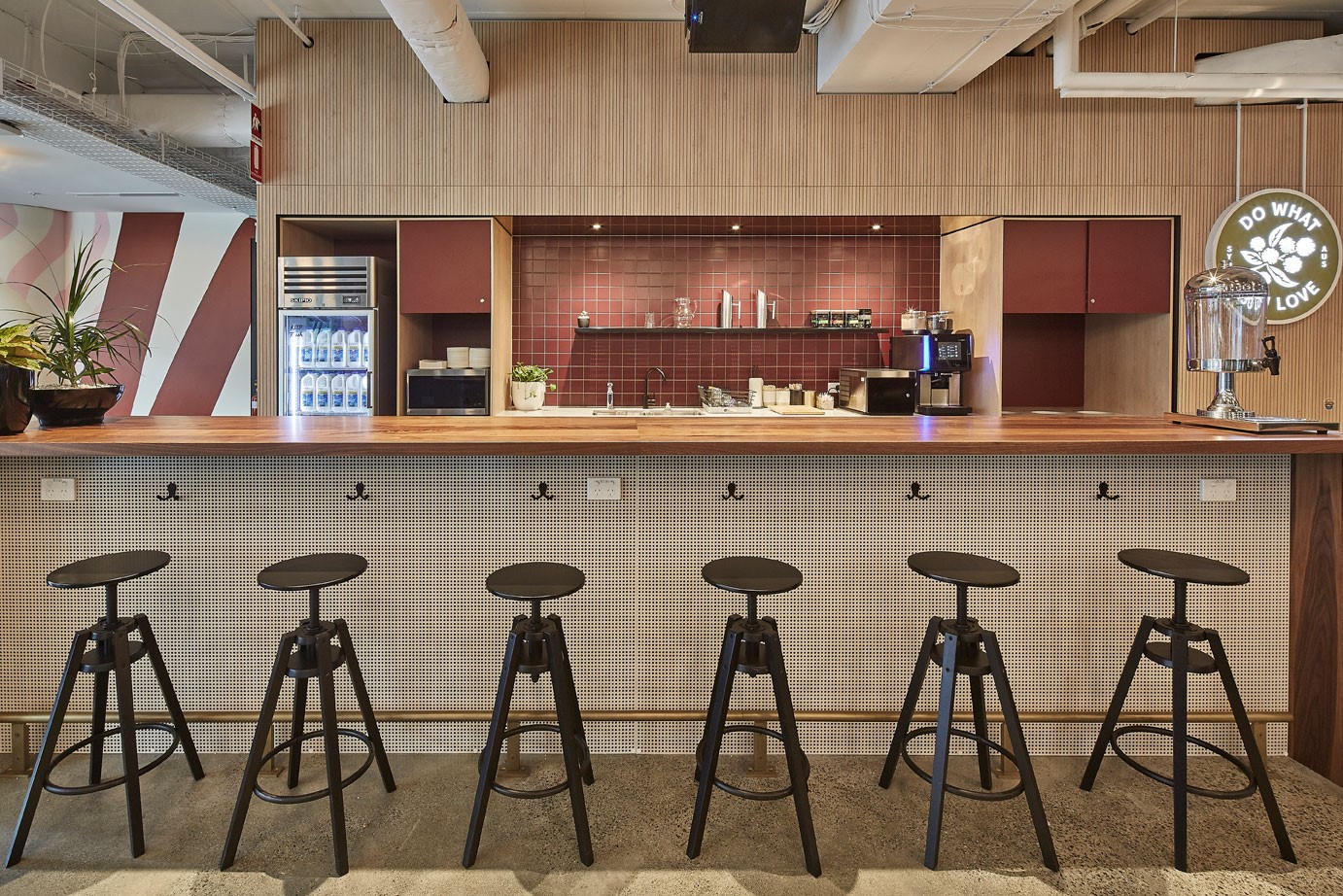 Modern office kitchenette featuring a long wooden counter with black bar stools and a tiled backsplash.
