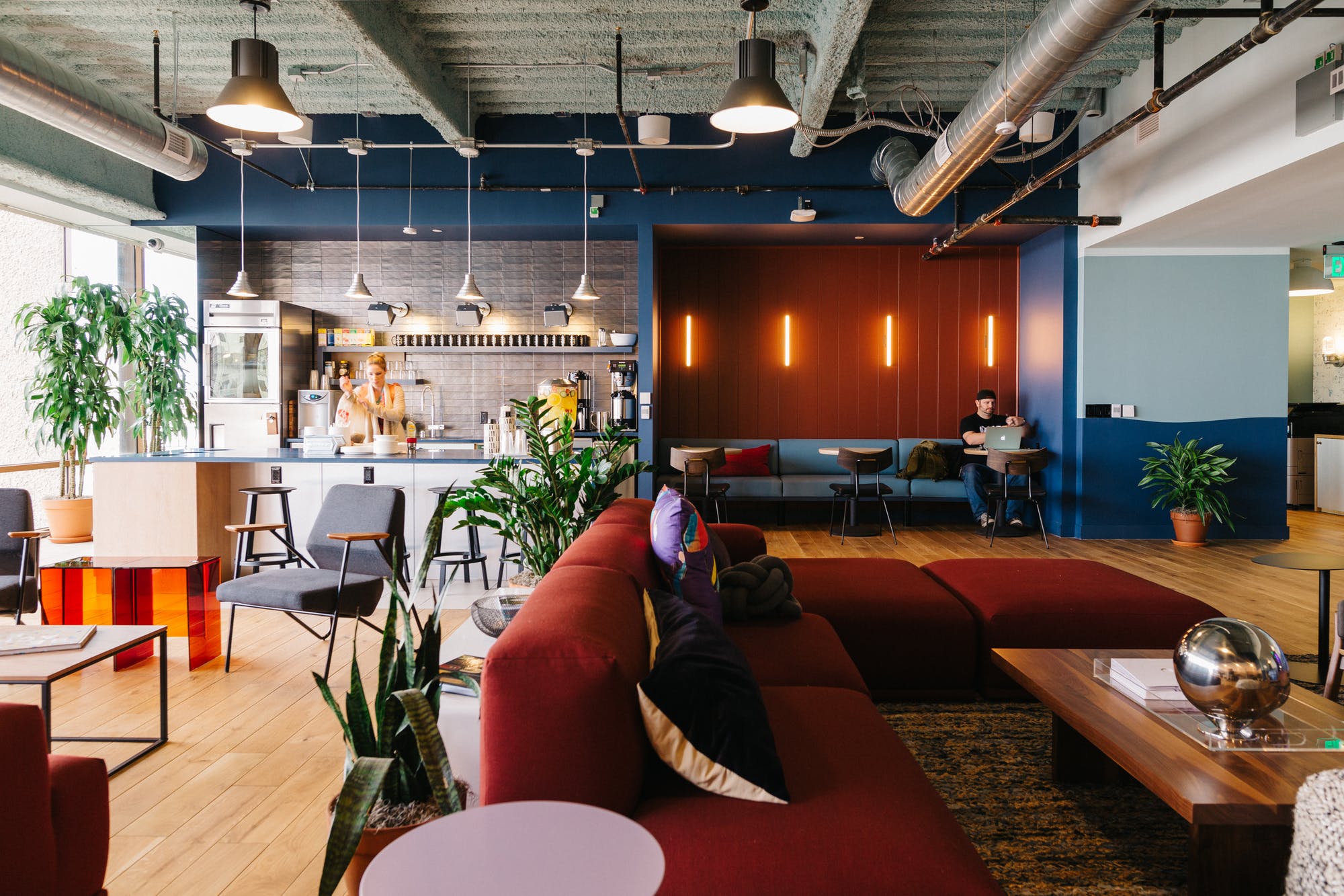 Modern office lounge featuring a red sofa, a kitchenette, and a seating area with warm wall lighting.