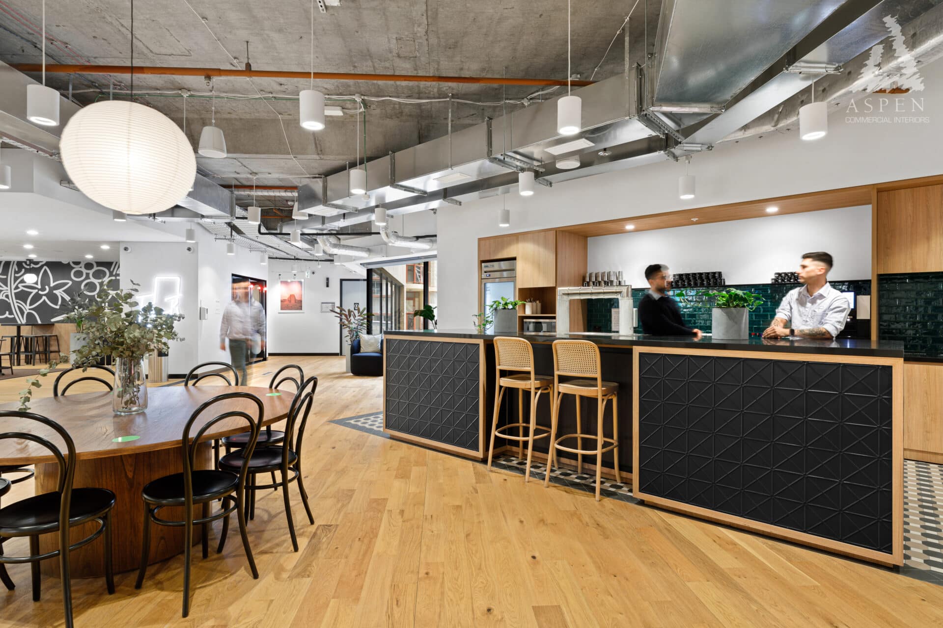 Modern open-plan office featuring a wooden dining table, a long black service counter, and industrial concrete ceilings.