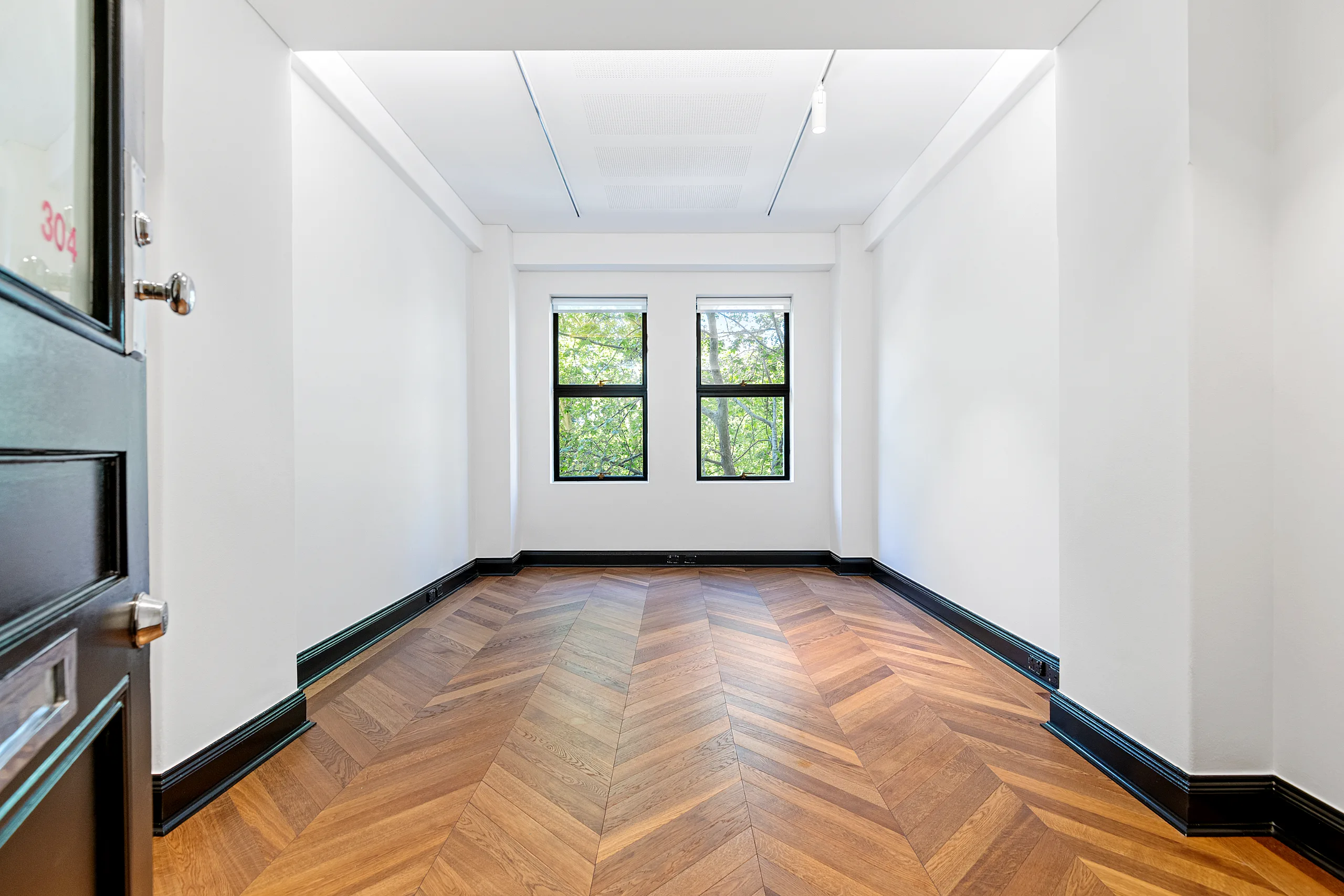 Empty office room with white walls, herringbone timber flooring, and two windows overlooking green trees.