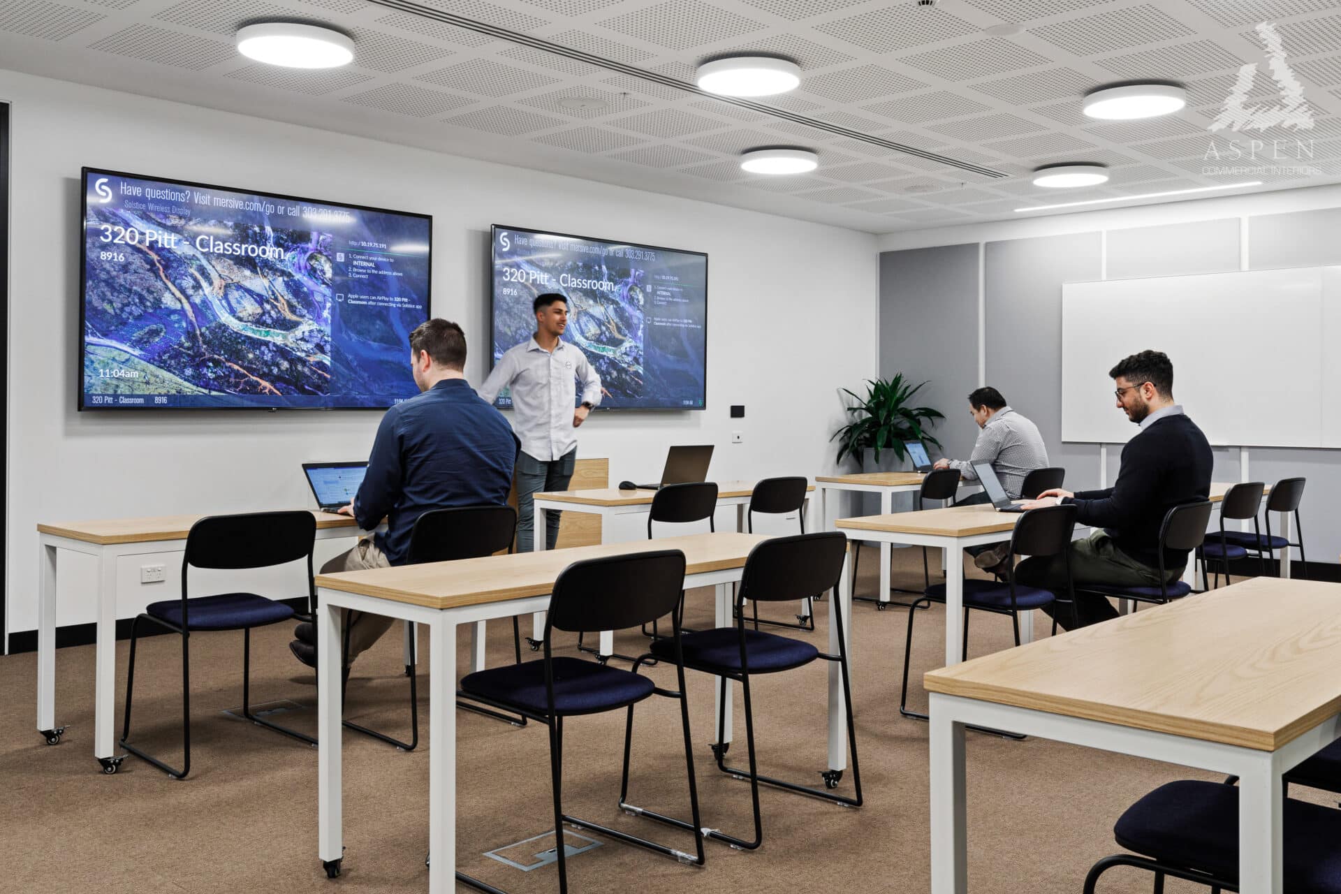 Bright classroom with long wooden desks, black chairs, and a large presentation screen on a white wall.