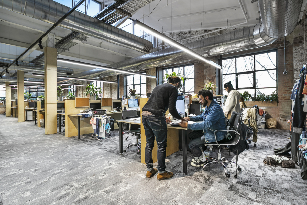 Bright, industrial-style open plan office with large windows, exposed ductwork, and people working at wooden desks.