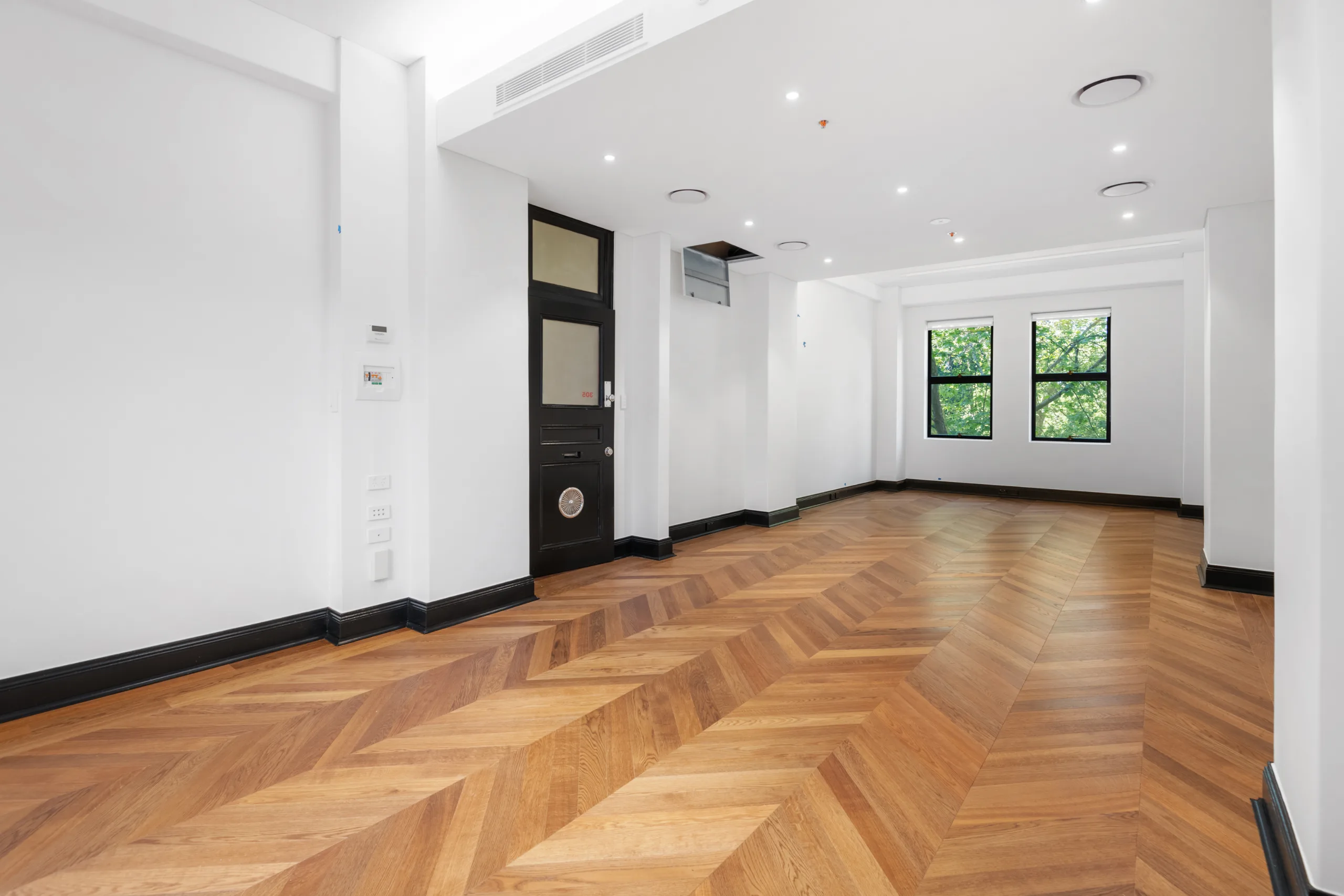 Empty office space with herringbone timber flooring, white walls, and large windows overlooking green trees.