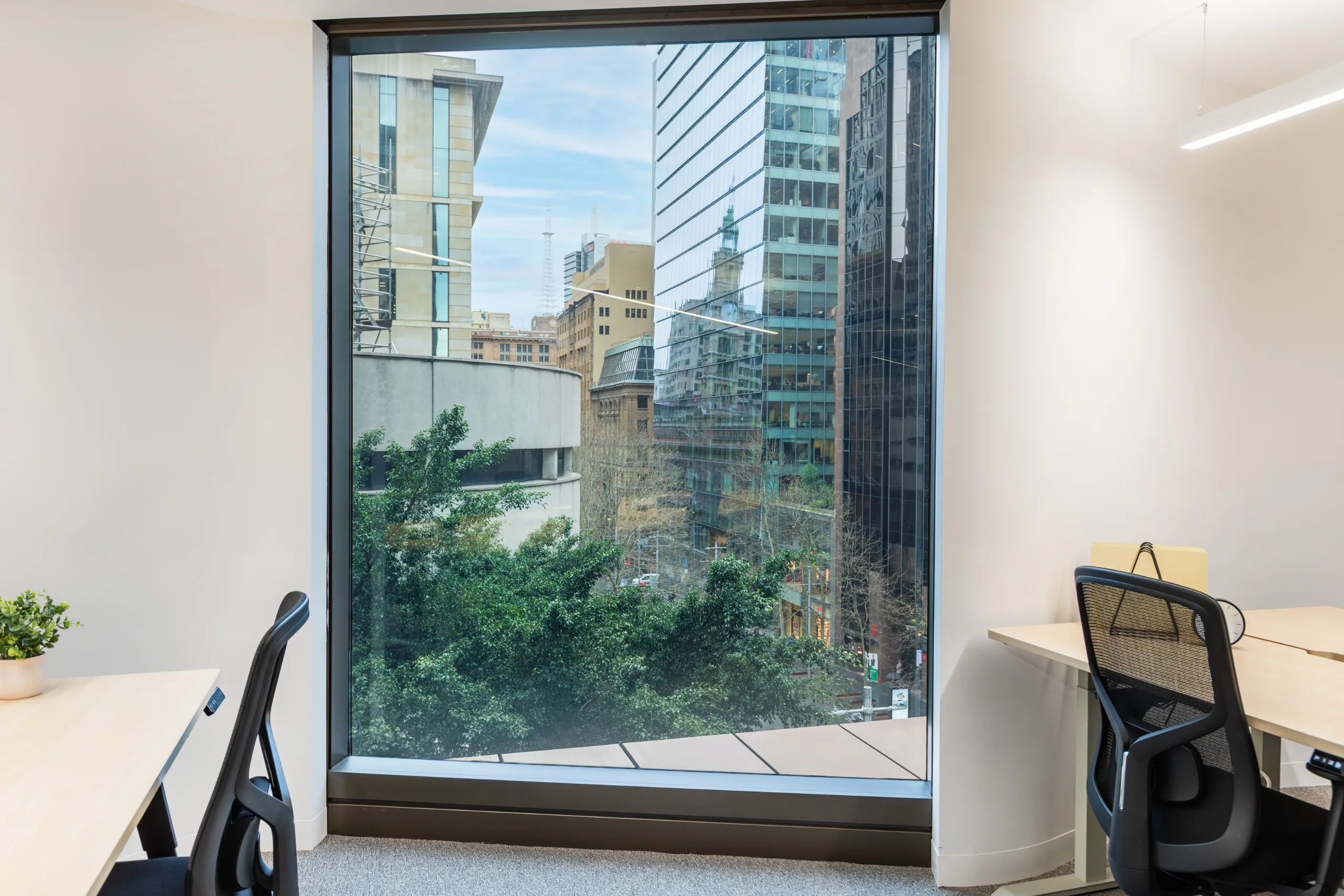 Bright office space with desks, ergonomic chairs, and a large window overlooking city buildings and greenery.