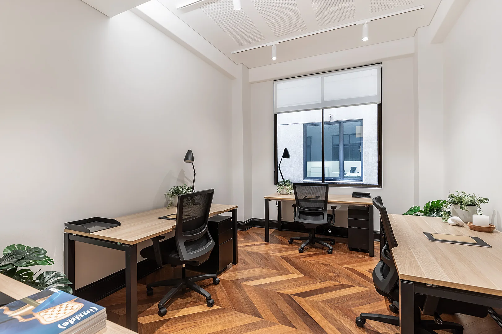 Brightly lit office with wooden desks, black chairs, and large windows overlooking a city building.