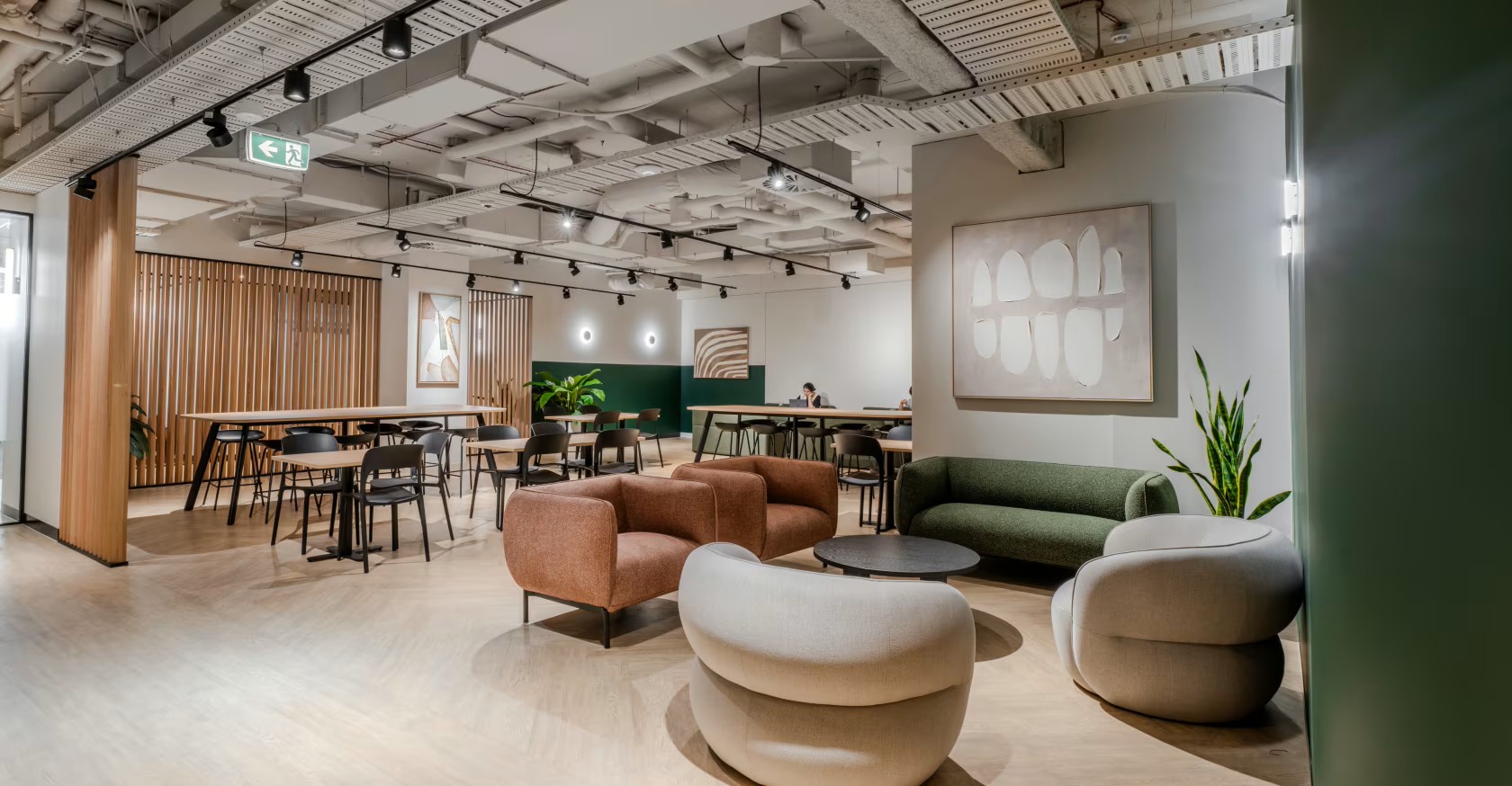 Modern open-plan office lounge featuring comfortable armchairs, a green sofa, and long communal desks under bright lighting.