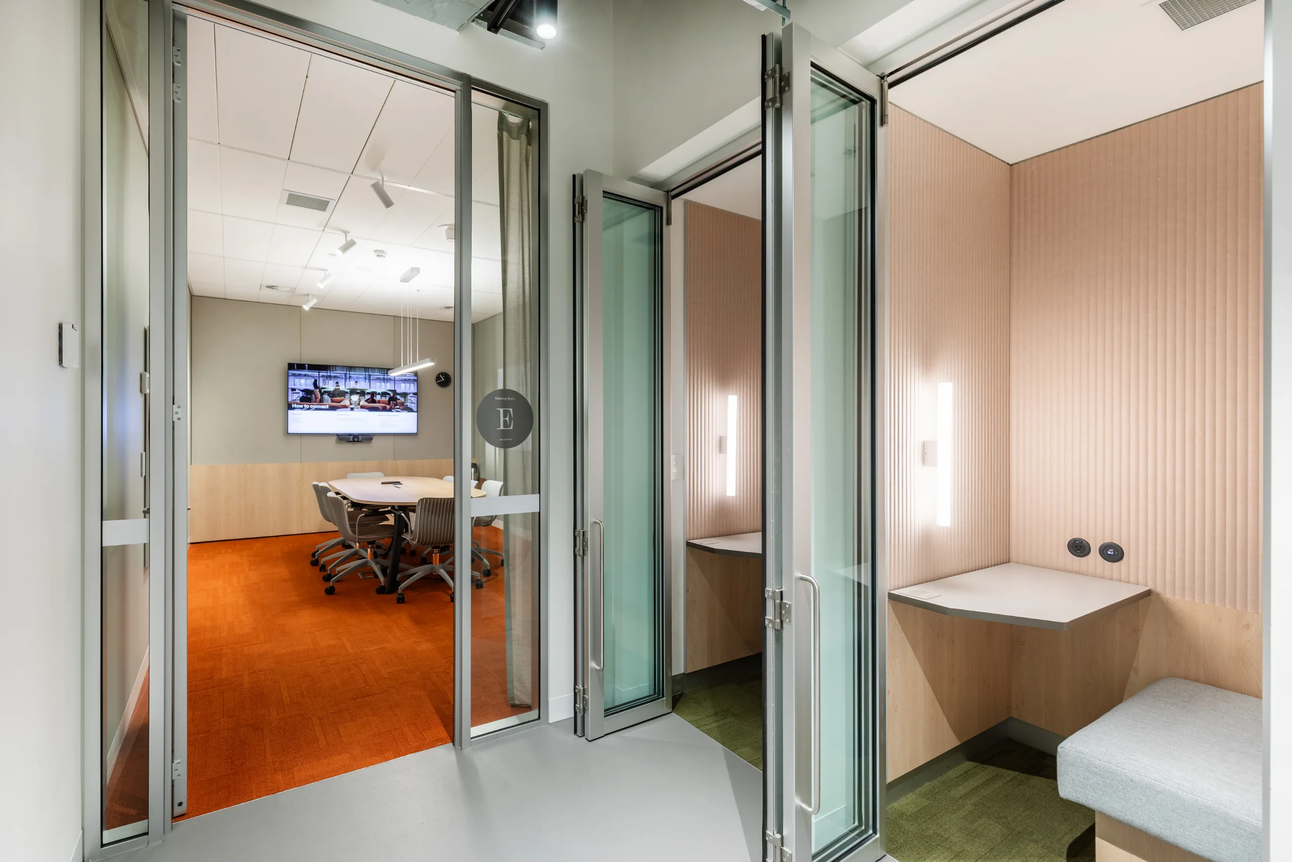 Glass-walled meeting rooms with bright lighting and modern furniture overlook a corridor in a flexible office space.