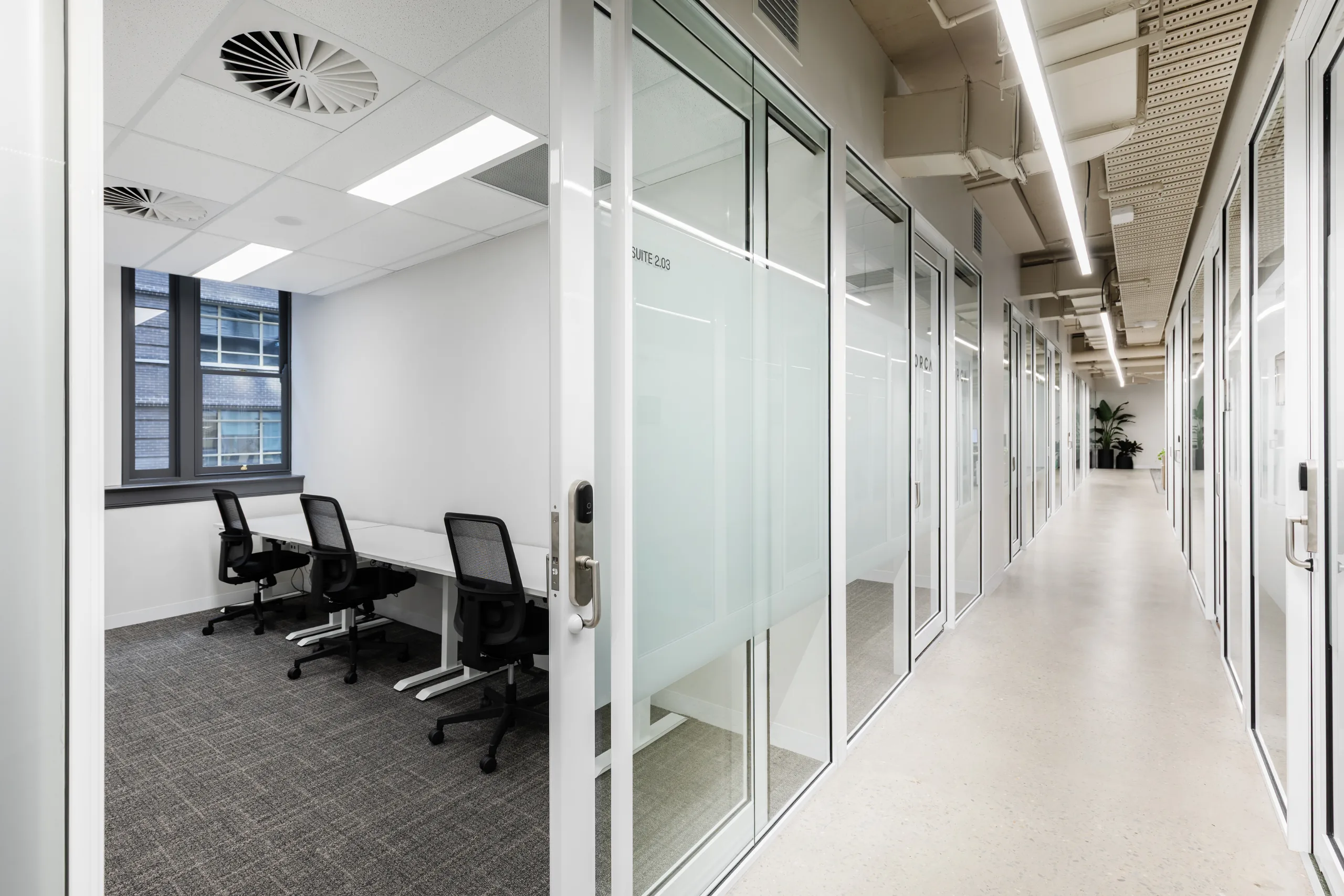 Bright office corridor with glass-walled private suites containing desks, chairs, and large windows.