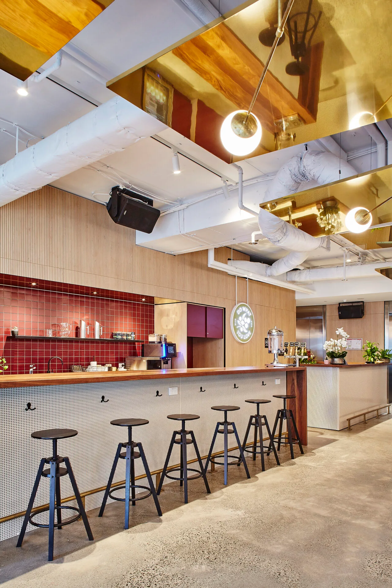 Modern office kitchen area with a long wooden bar, black stools, and red tiled walls under reflective ceilings.