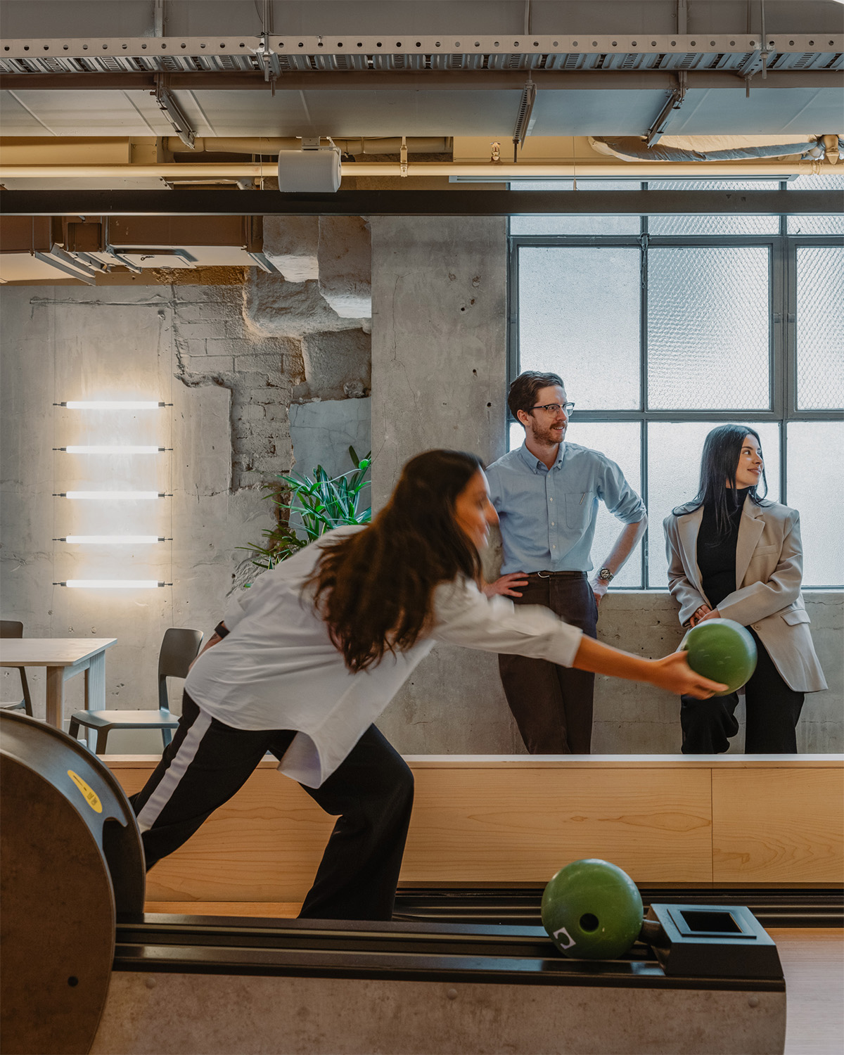 A person bowls a green ball in a bright, modern office space with large windows and concrete walls.