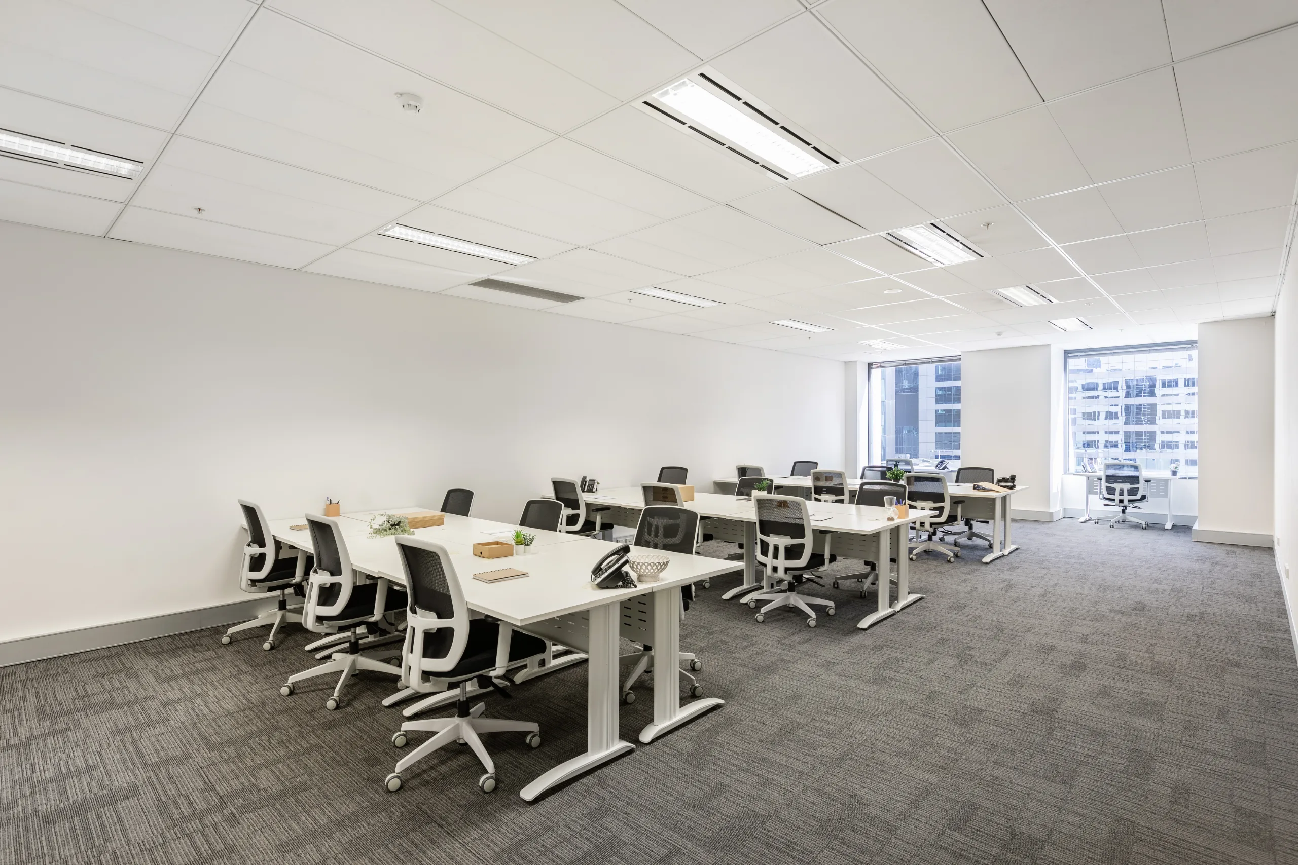 Bright, modern office space featuring long white desks, ergonomic chairs, and large windows with city views.