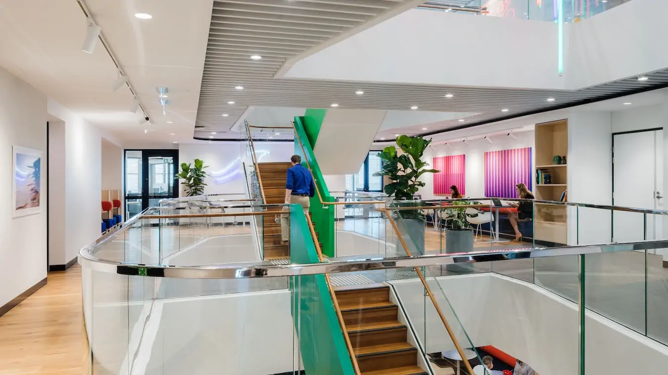 Bright, modern open-plan office featuring wooden stairs, glass railings, and large indoor plants under bright ceiling lights.