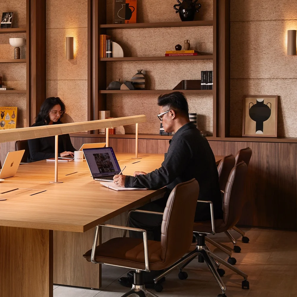 Two people work at a long wooden desk in a modern office with warm lighting and built-in shelving.