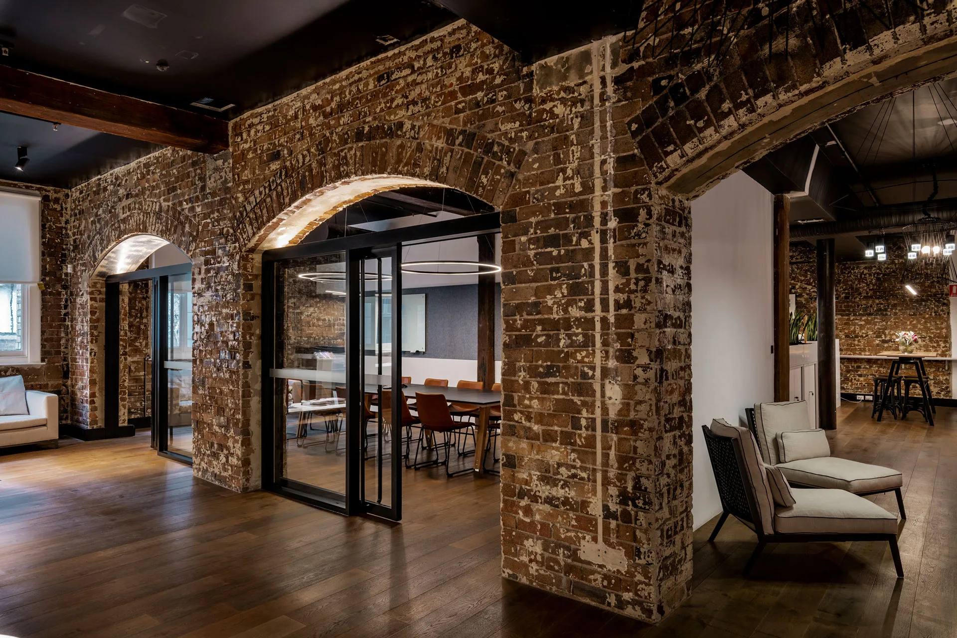 Modern office space featuring exposed brick arches, wooden floors, and a glass-walled meeting room with warm lighting.