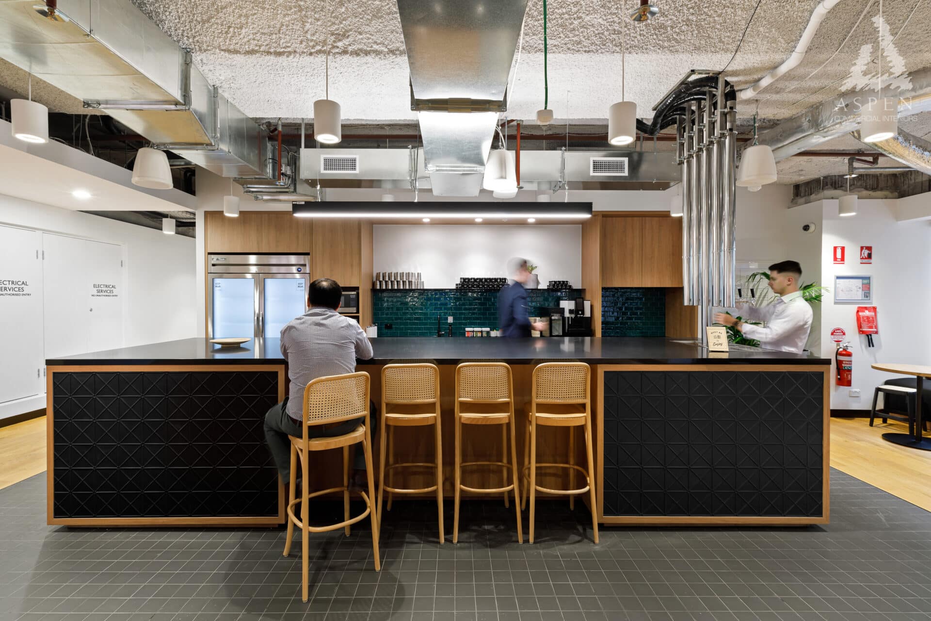 Modern office kitchen area with a long black breakfast bar, wooden stools, and bright overhead lighting.