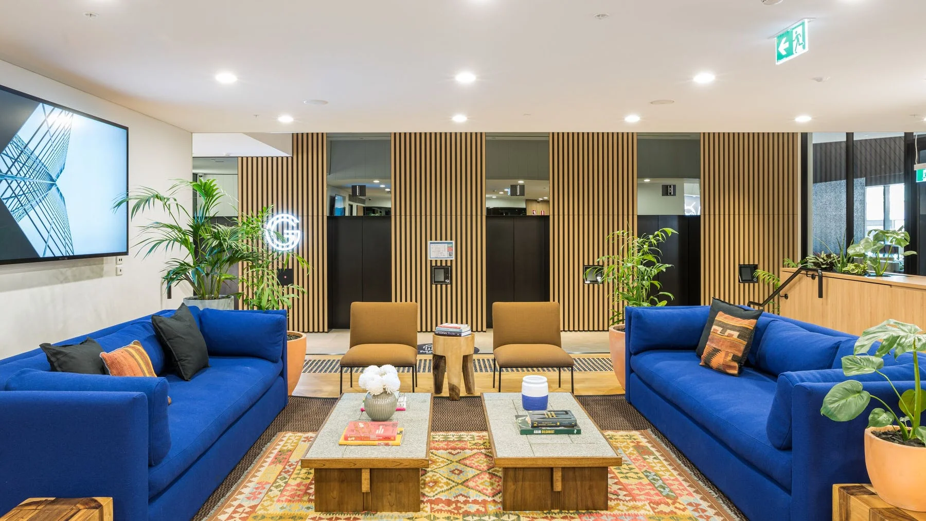 Modern lounge area with blue sofas, wooden coffee tables, and potted plants against a slatted timber wall.