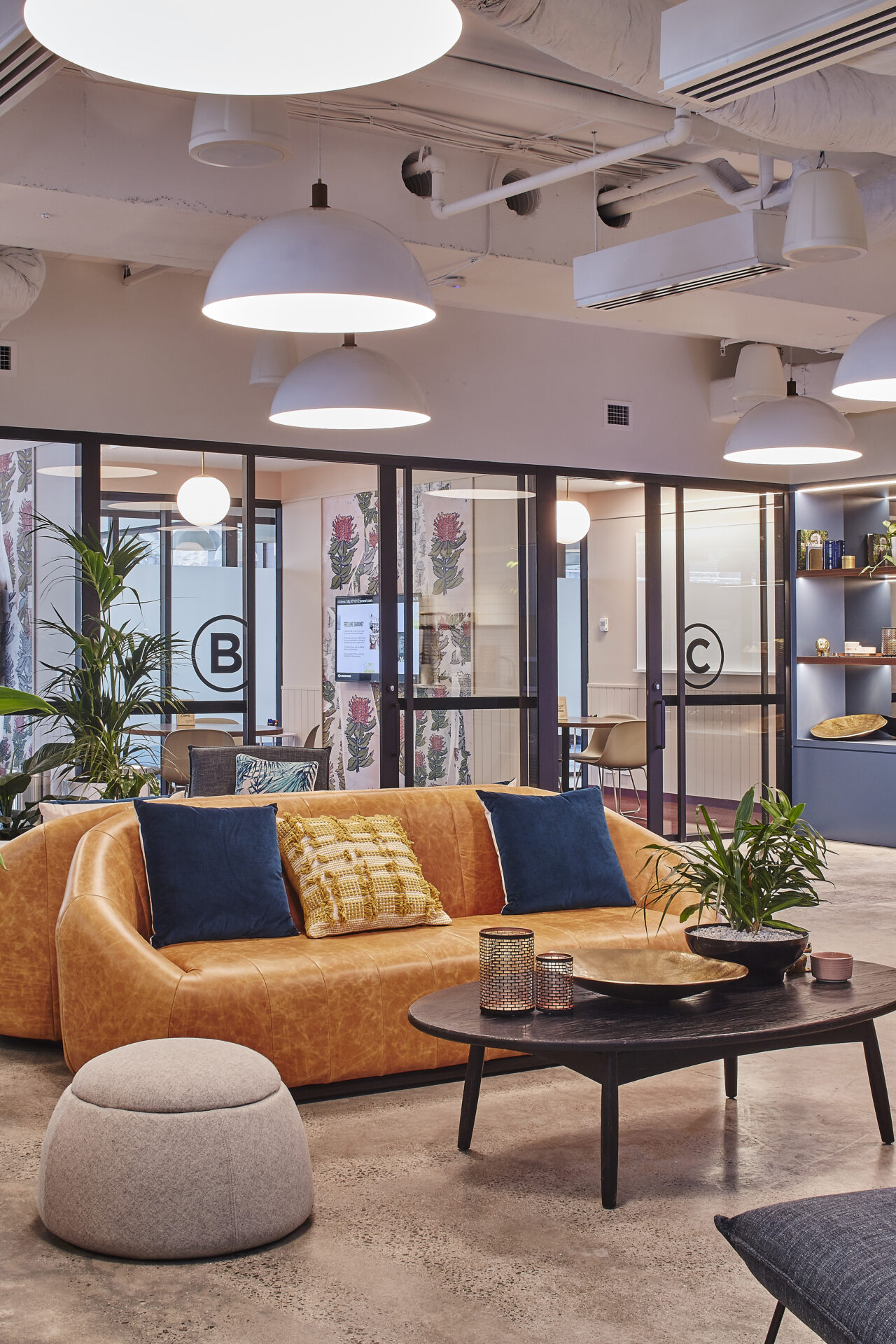 Modern lounge area with a tan leather sofa, patterned cushions, and large windows in a bright office space.