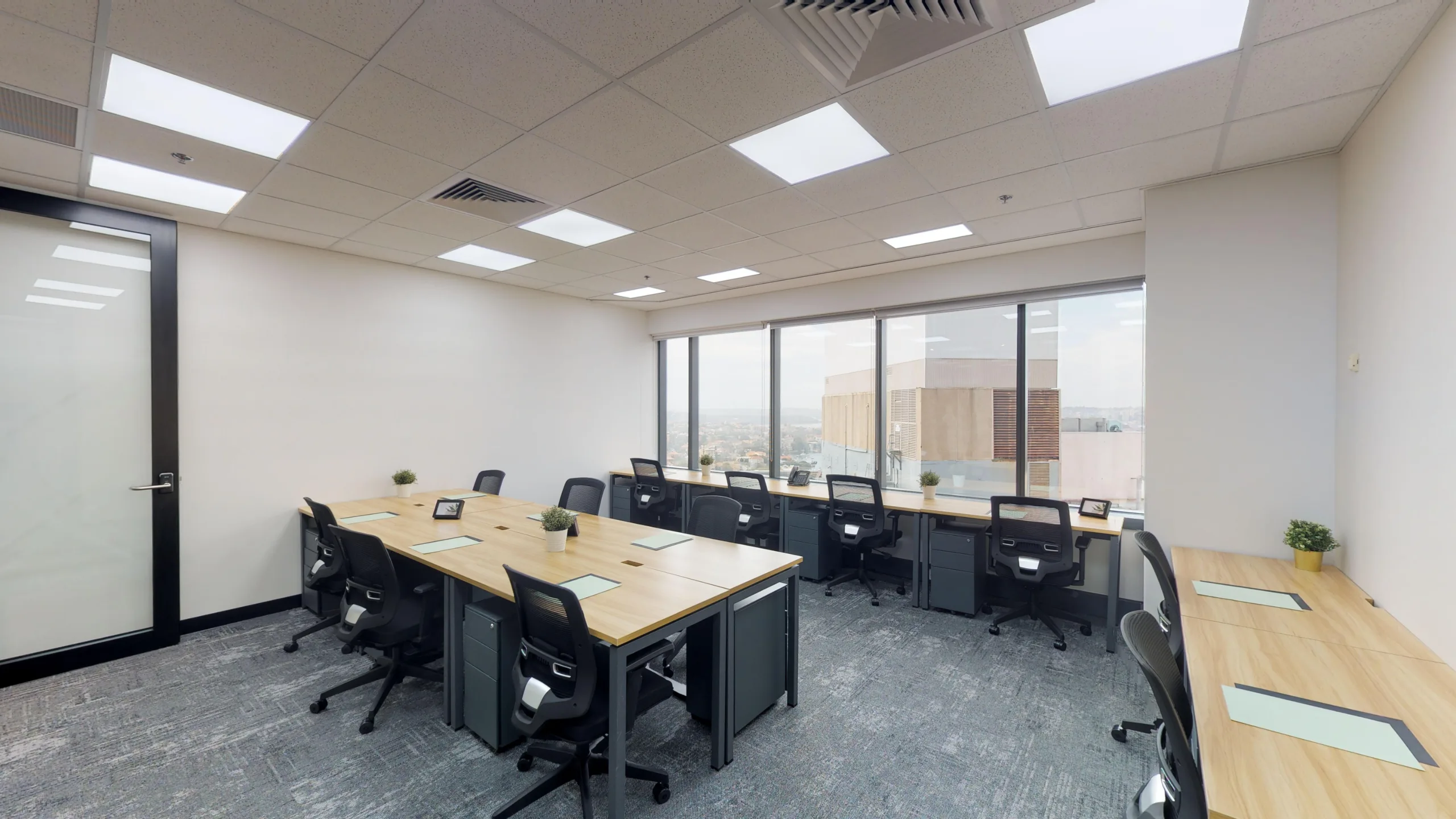 Bright, modern office space with wooden desks, black ergonomic chairs, and large windows overlooking a city view.