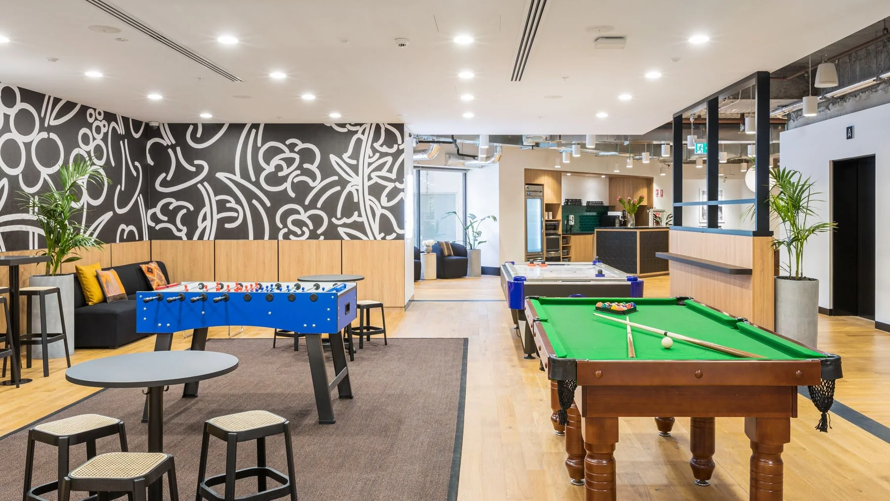 Brightly lit office breakout area featuring foosball, pool tables, and seating on light timber flooring.