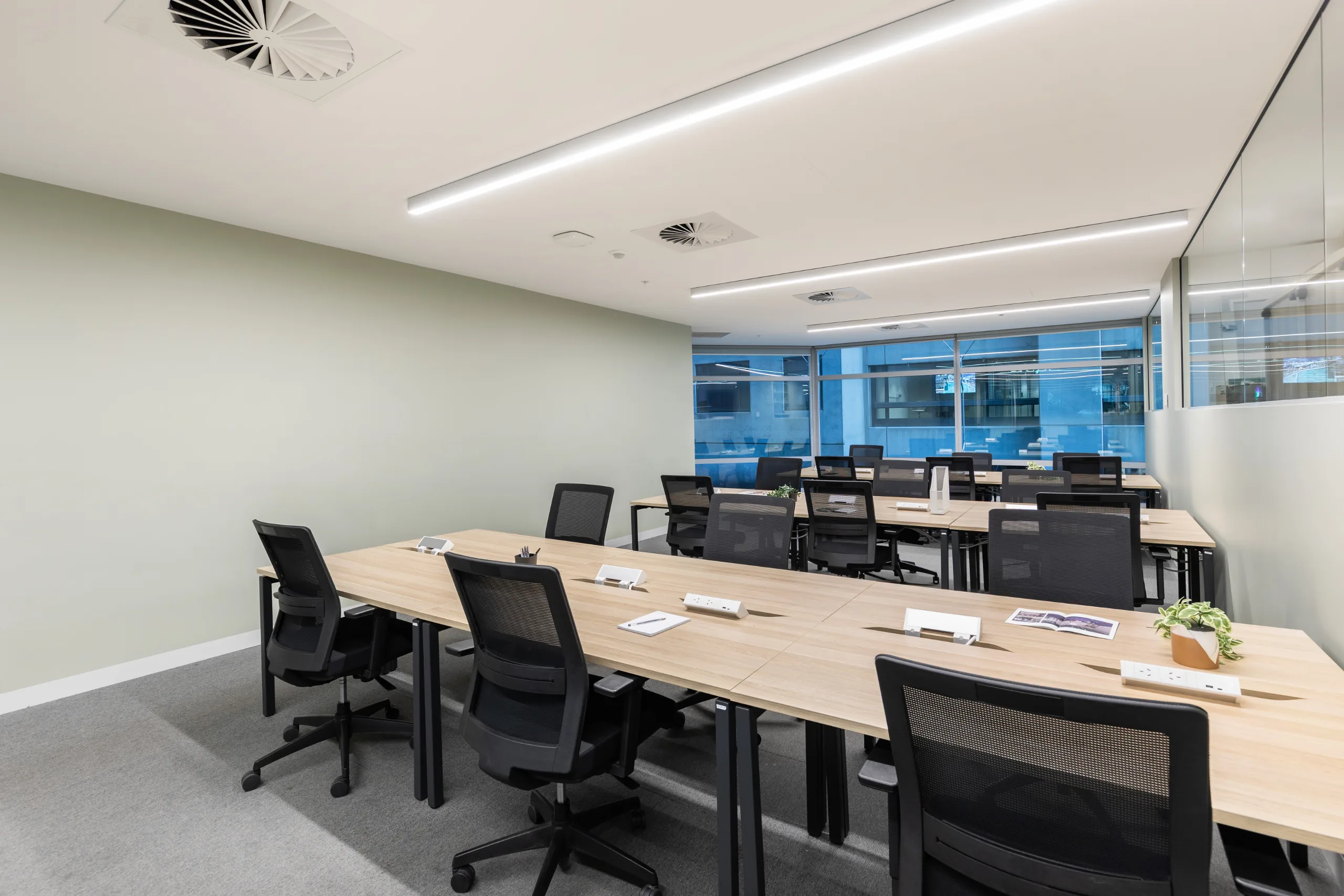Brightly lit meeting room featuring long wooden desks, black mesh chairs, and large windows with city views.