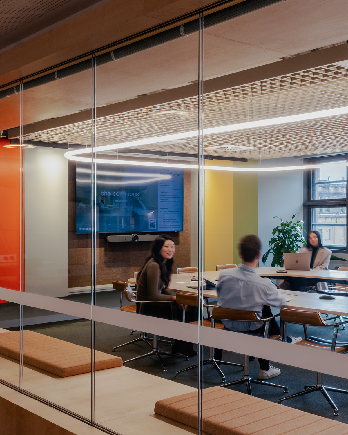 Brightly lit meeting room with a large screen, wooden table, and people sitting in modern chairs.