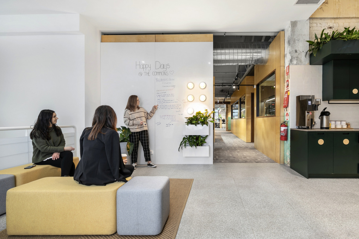 Two people sit on modular lounge seating near a large whiteboard in a bright, modern office common area.