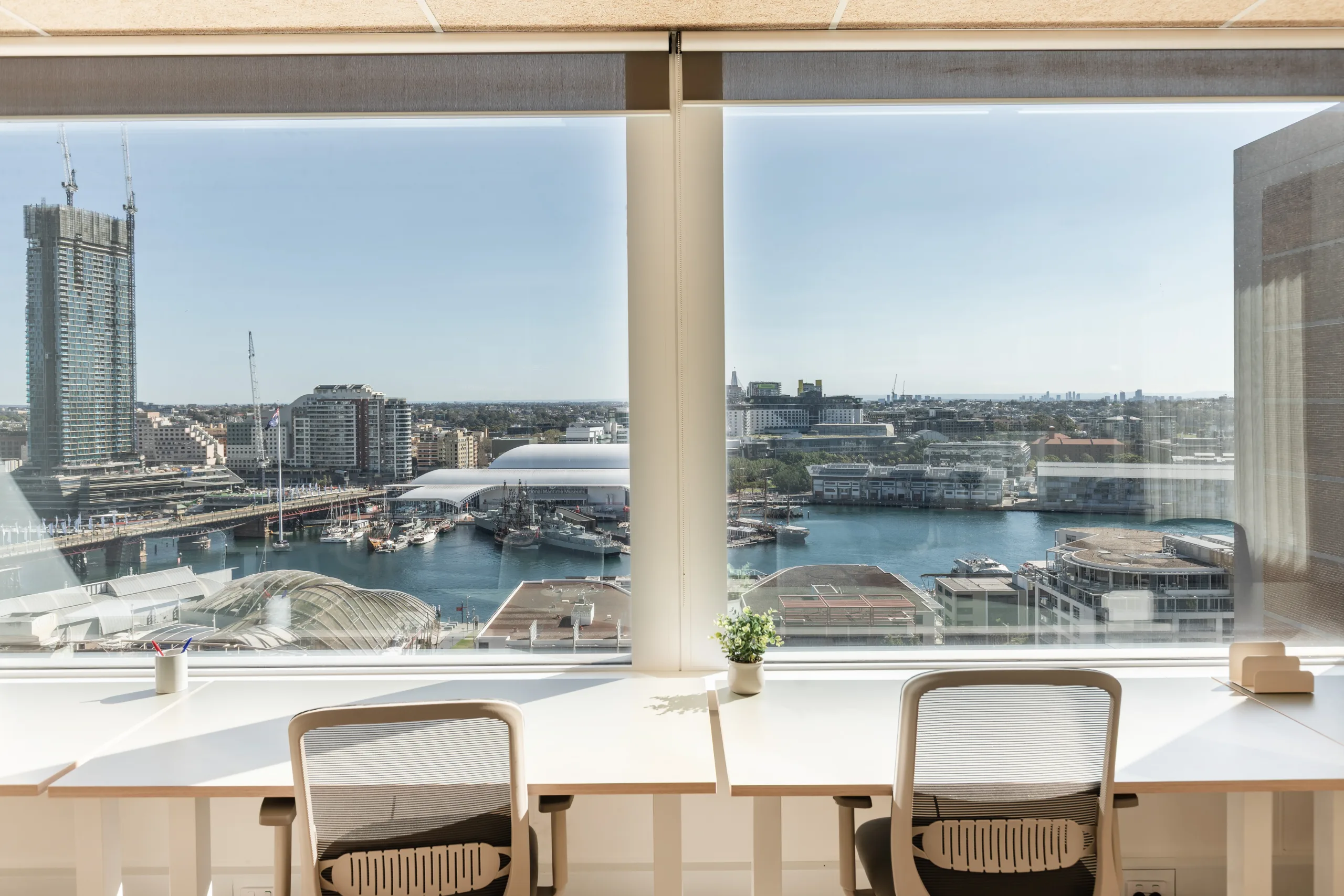 Bright office workspace with white desks and chairs overlooking a sunny harbour and city skyline.