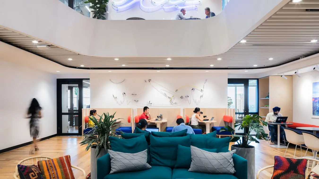 Bright, modern office lounge with teal sofas, wooden floors, and people working in a sunlit open space.