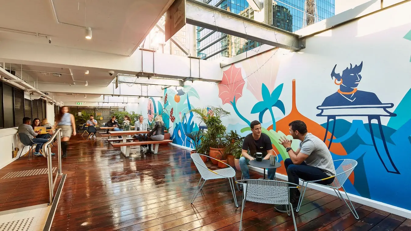 Bright outdoor terrace with wooden decking, colourful murals, and people sitting at tables in natural light.